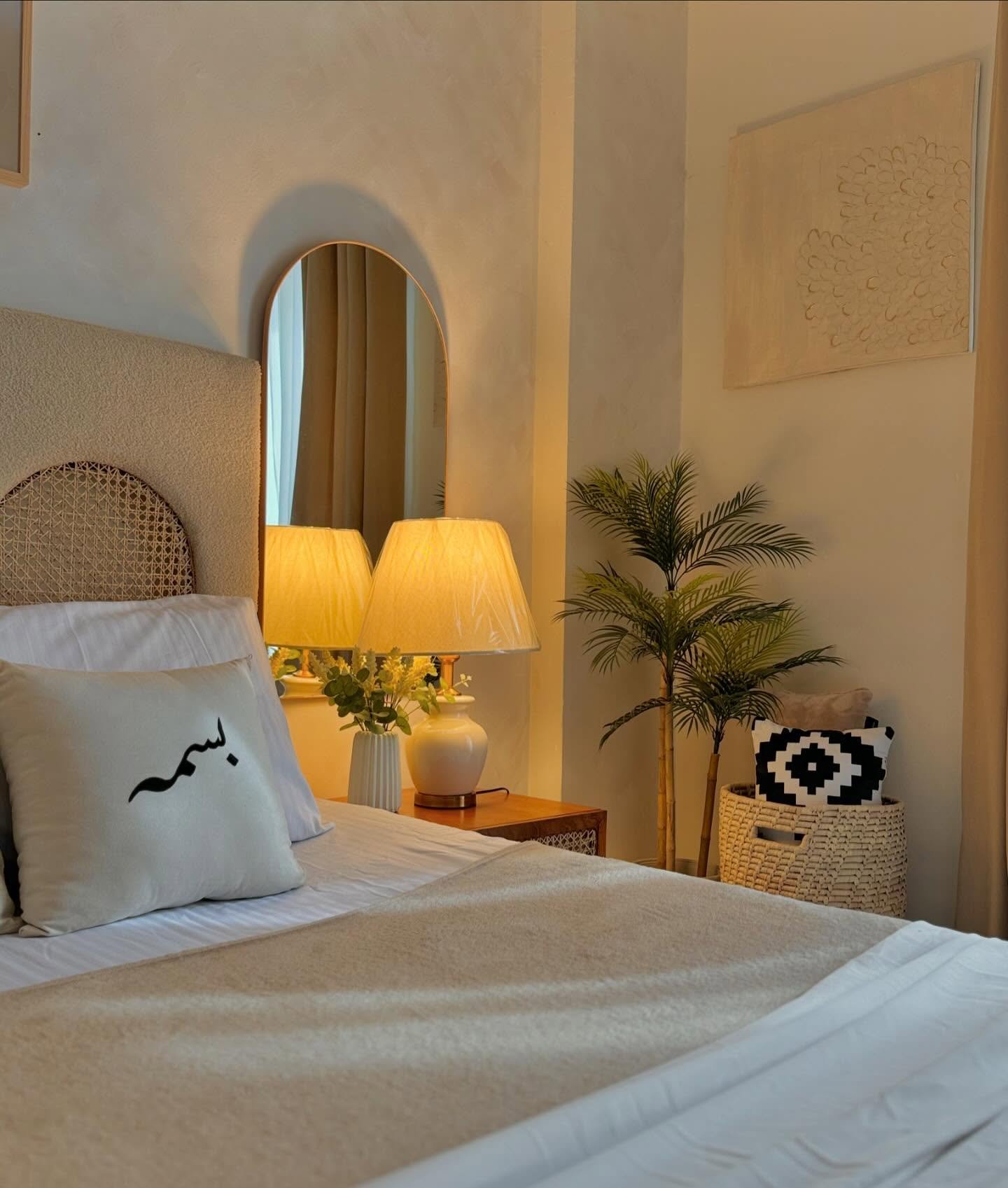 A close-up of a neutral-toned bedroom with a lamp on a wooden nightstand. The lamp emits a warm, soft glow.
