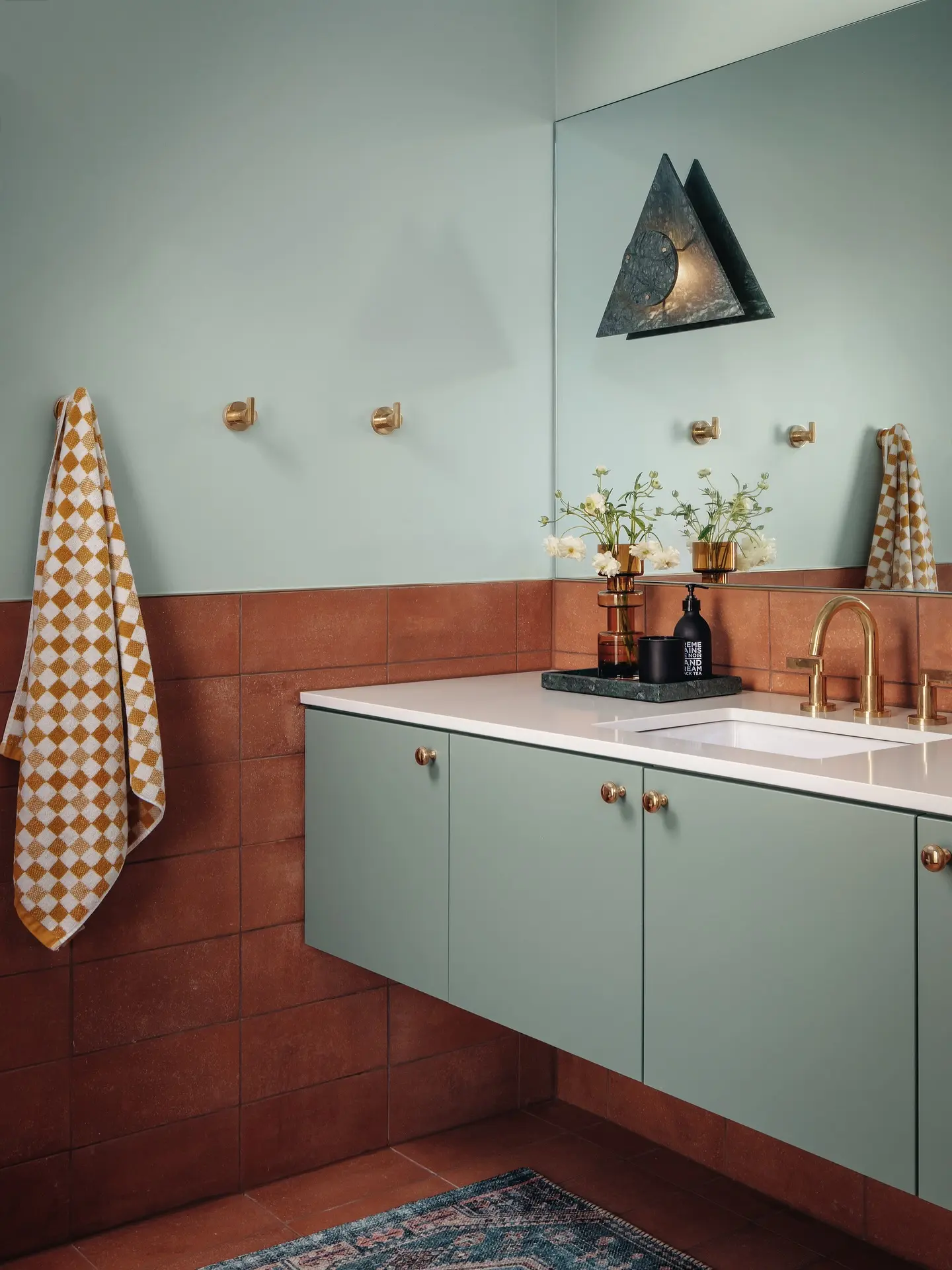 A floating, pale green bathroom vanity with a white countertop and gold fixtures, set against a two-tone wall with dark terracotta-colored tiles on the bottom and light green paint on top.