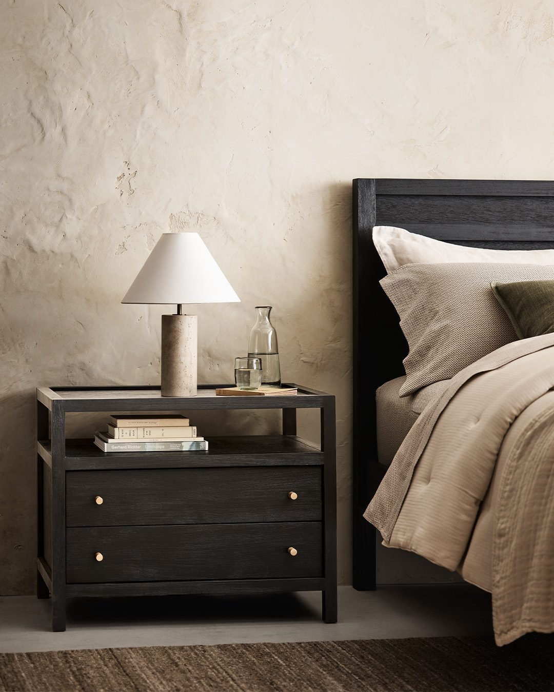 A dark, almost black, two-drawer nightstand with brass knobs. The top shelf holds a textured concrete lamp with a white shade, a glass carafe, a small glass, and a couple of books. The wall behind is a light, textured plaster finish.