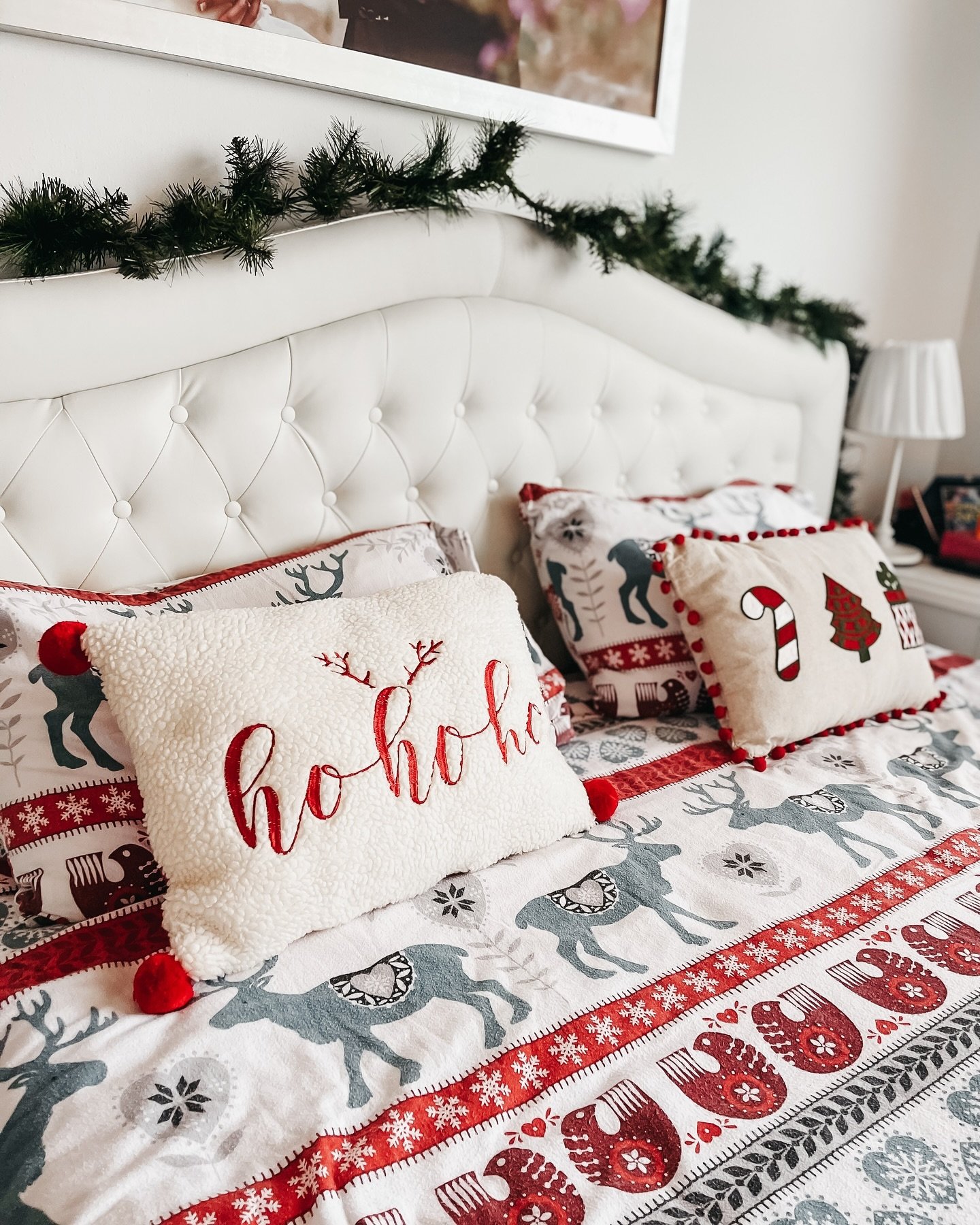 Close-up of a white, tufted headboard draped with a simple pine garland, and a bed featuring festive red, white, and gray Nordic-style reindeer bedding.