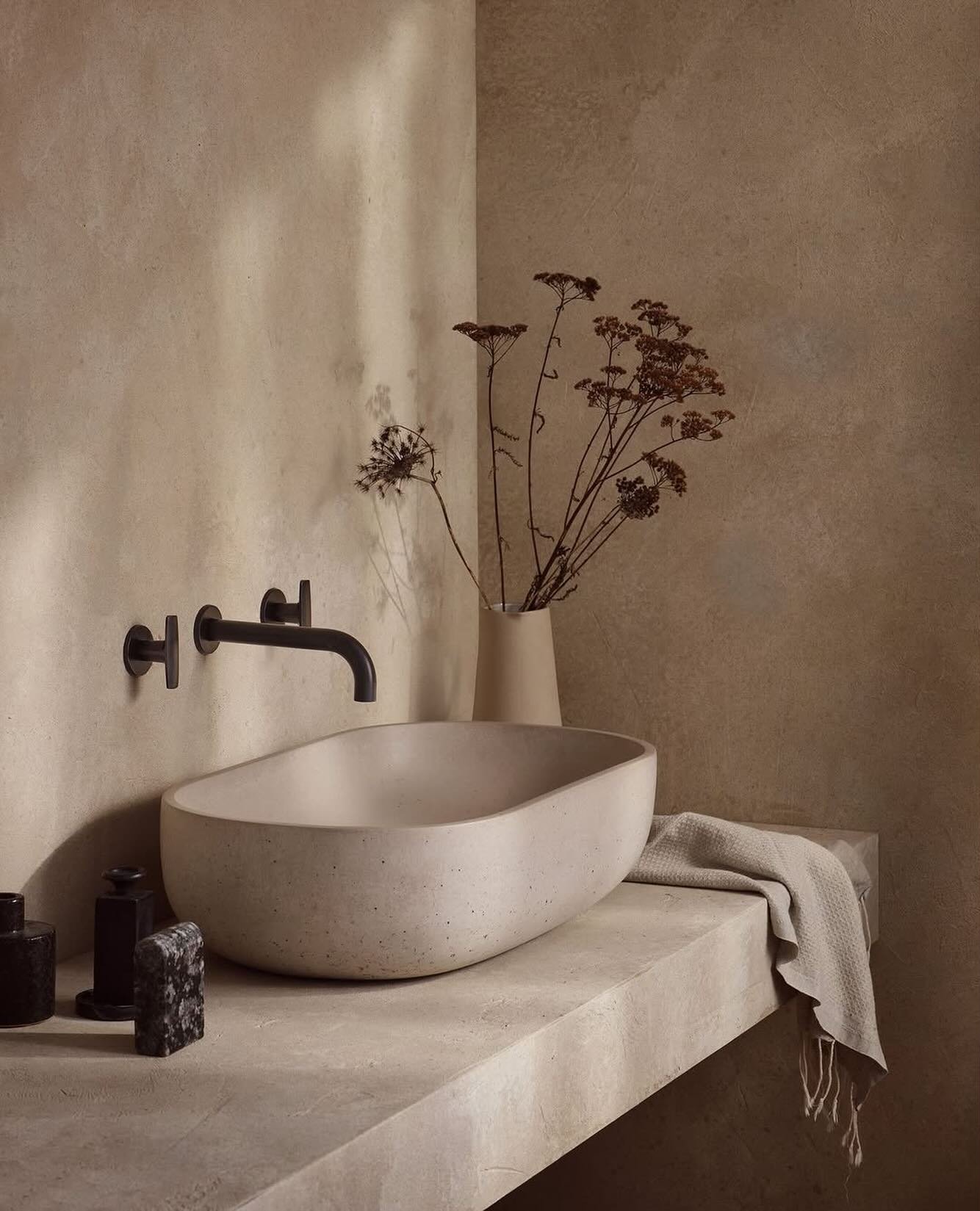 A minimalist, rustic bathroom with a large, oval-shaped concrete vessel sink. The sink rests on a textured concrete countertop against a warm, beige plaster wall, complemented by a matte black wall-mounted faucet and dried flowers in a vase.