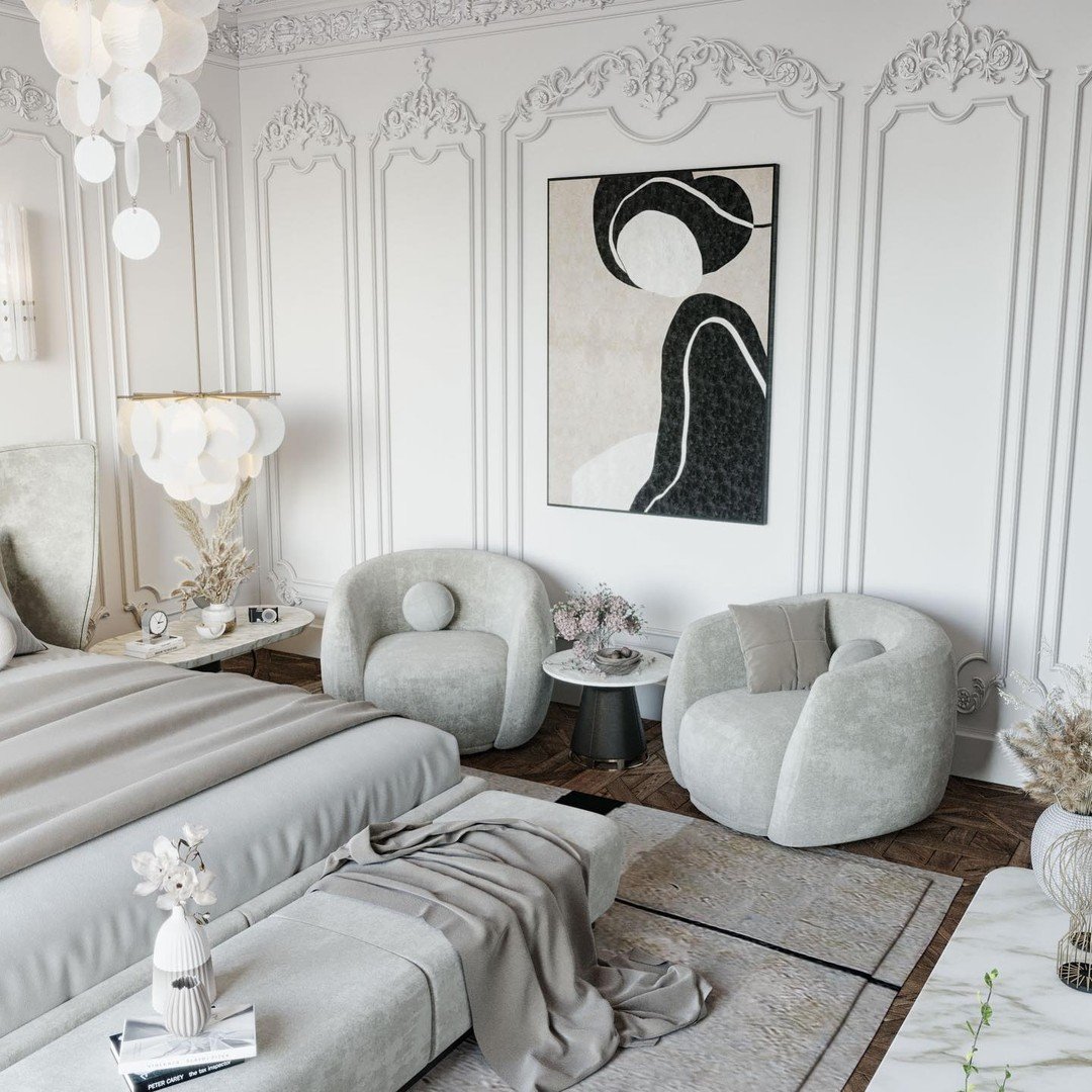 A pair of modern bouclé armchairs flank a small round table at the foot of a bed in a luxurious white bedroom.
