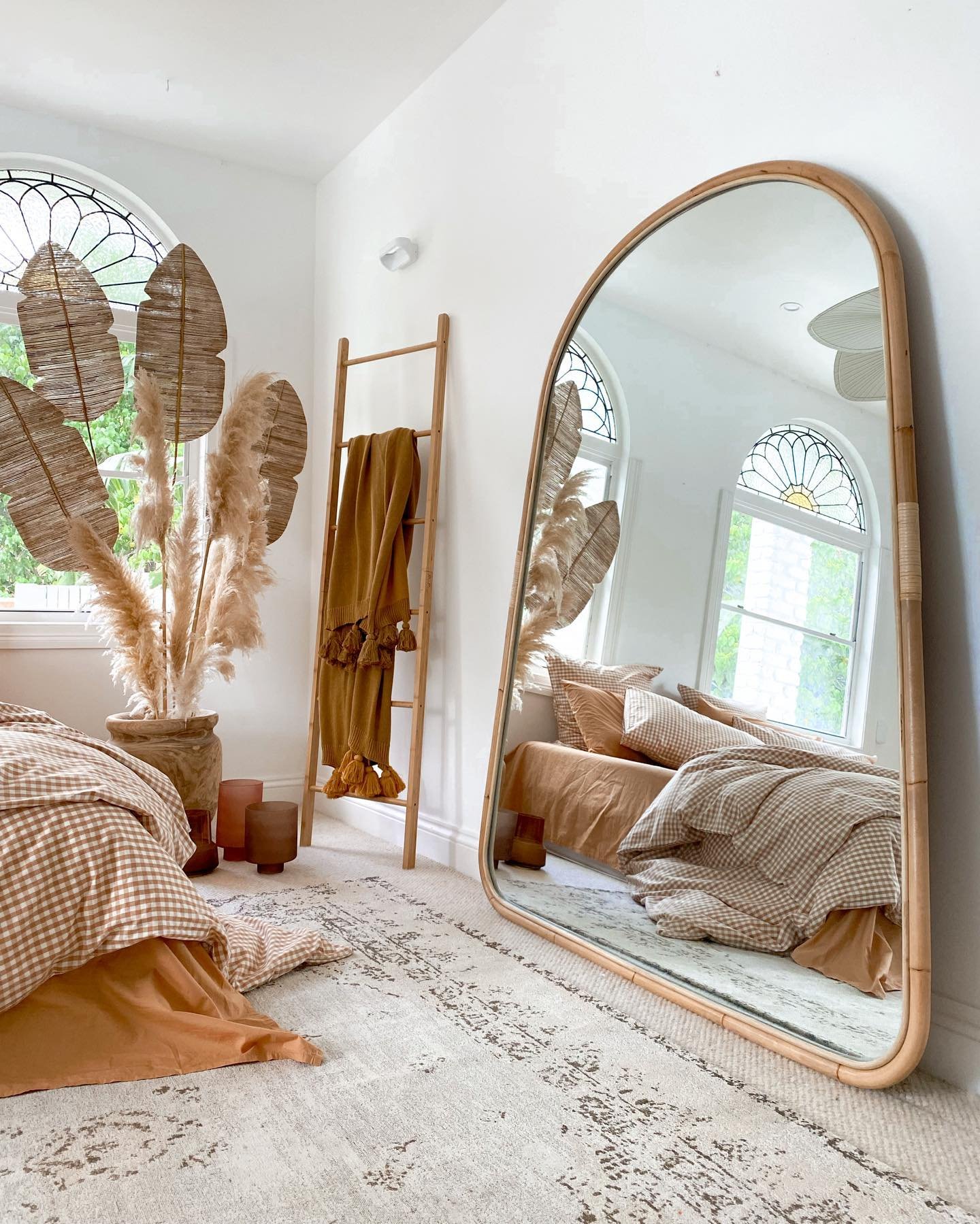 A tall, arched, full-length mirror with a light wood frame leans against a white bedroom wall, reflecting a bed with earthy-toned gingham bedding and dried plants.