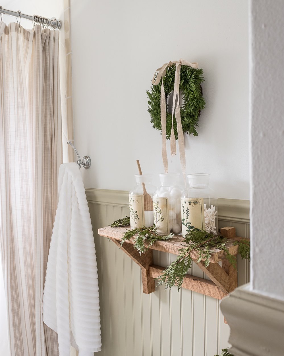 A wooden floating shelf in a bathroom decorated with draping pine garland, holding clear jars, and a small boxwood wreath hangs above with a burlap ribbon.