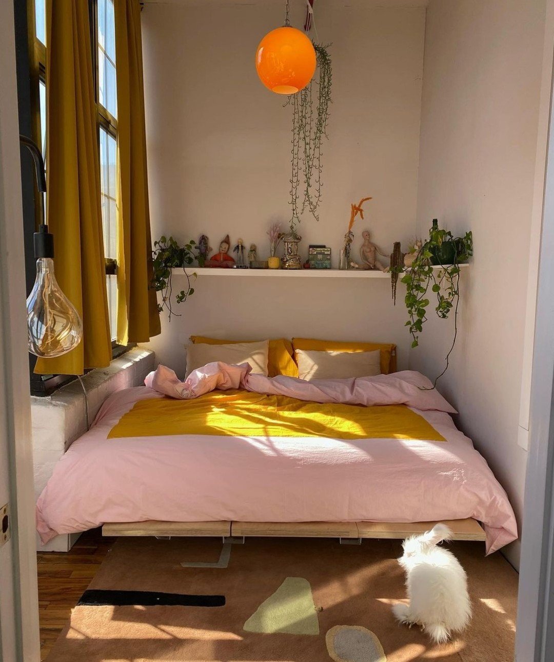 A bright bedroom with a bed featuring pink and yellow bedding, an orange spherical light fixture, and a floating shelf with plants and various figurines.
