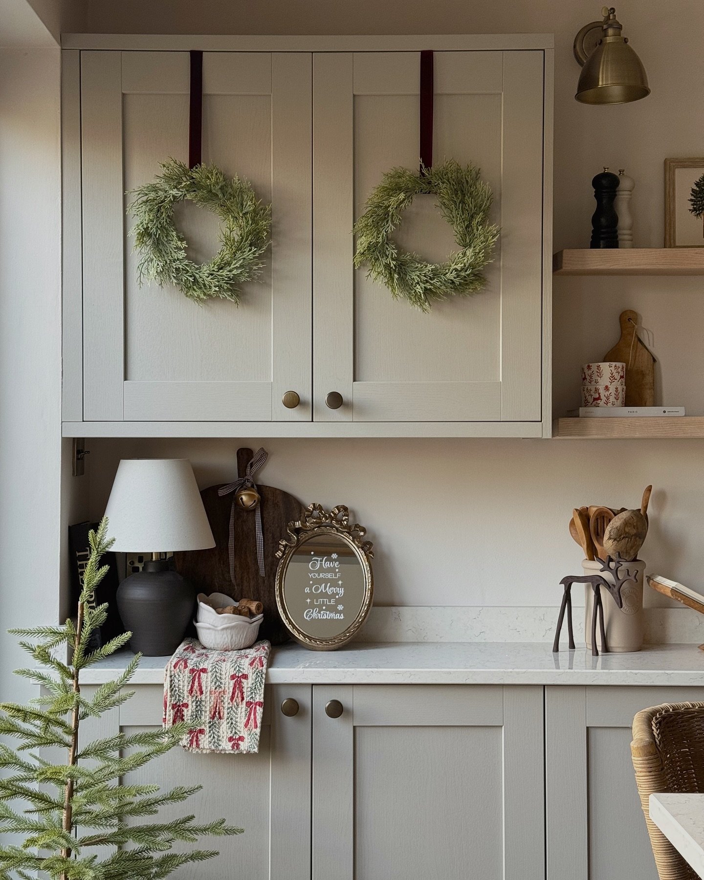 A close-up of a cozy, neutral kitchen with sage green shaker cabinets. Two simple green wreaths hang on the upper cabinet doors with deep red ribbons, flanking a counter display featuring a small lamp, a gilded framed sign, and a miniature Christmas tree.