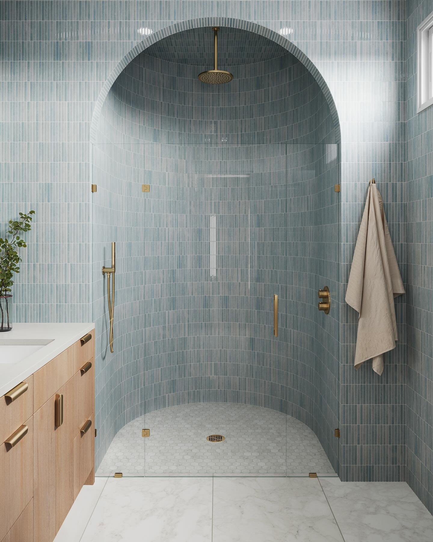 A modern bathroom featuring an arched walk-in shower with light blue vertical subway tiles and a small hexagonal tiled floor, a light wood vanity, and gold hardware.