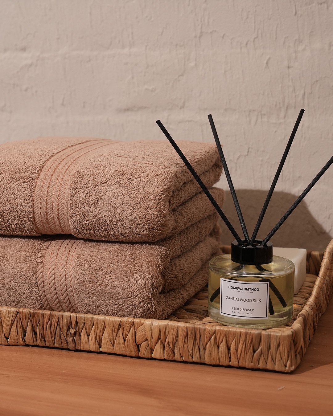 Two folded taupe towels displayed on a woven wicker tray next to a sandalwood silk reed diffuser.