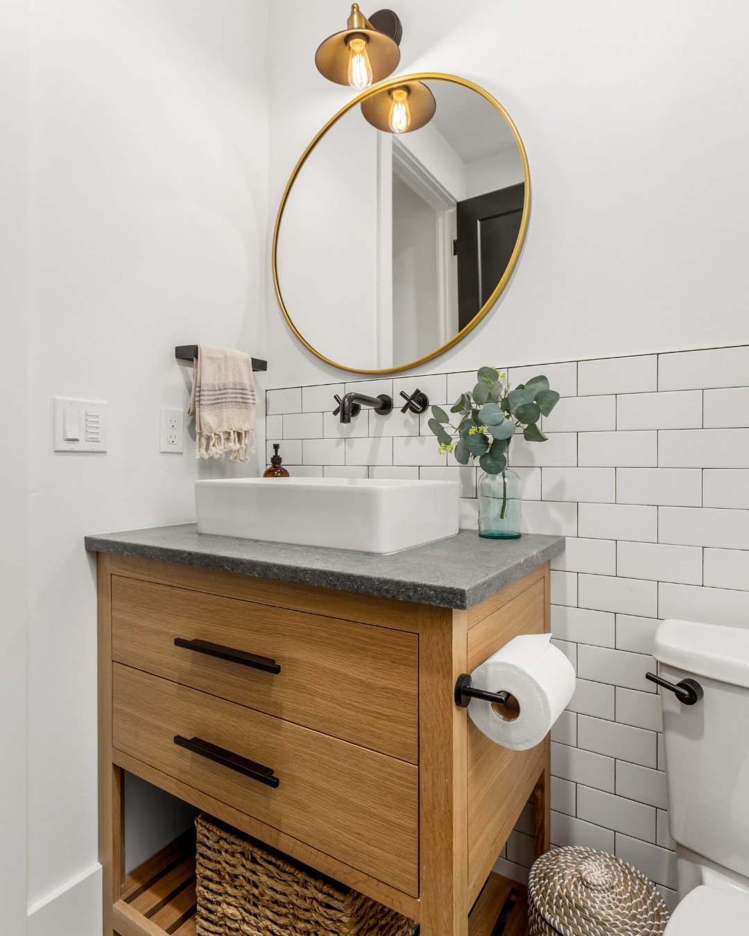 Natural wood vanity with a dark stone countertop and vessel sink. It features two drawers and an open bottom shelf holding a large woven storage basket.