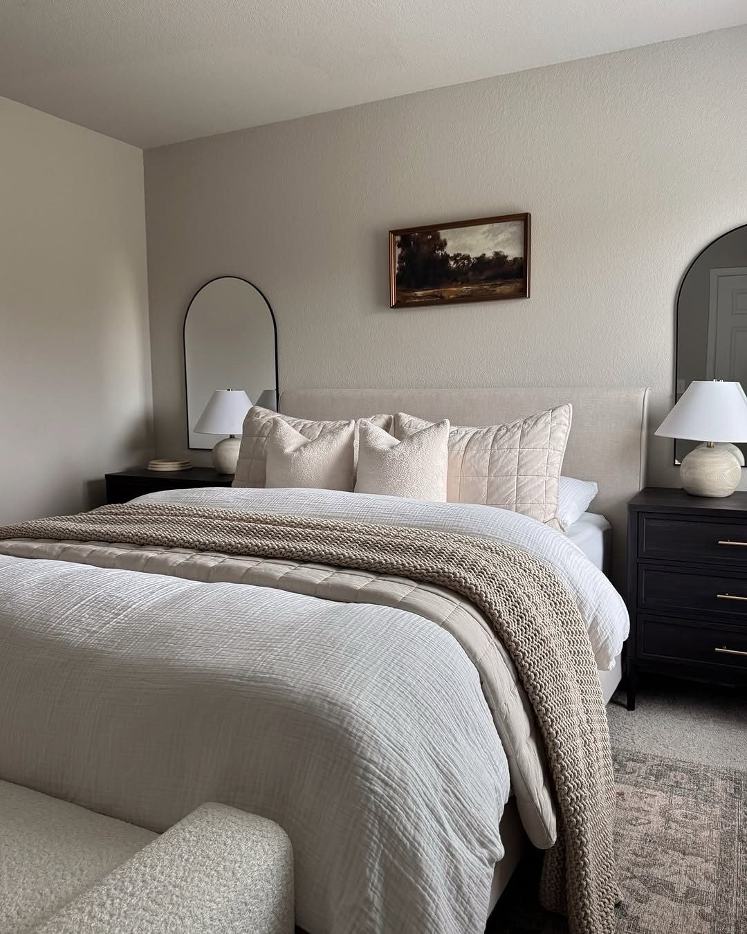 A cozy bedroom with a large bed dressed in a variety of cream and white textures, flanked by black nightstands with arched mirrors.