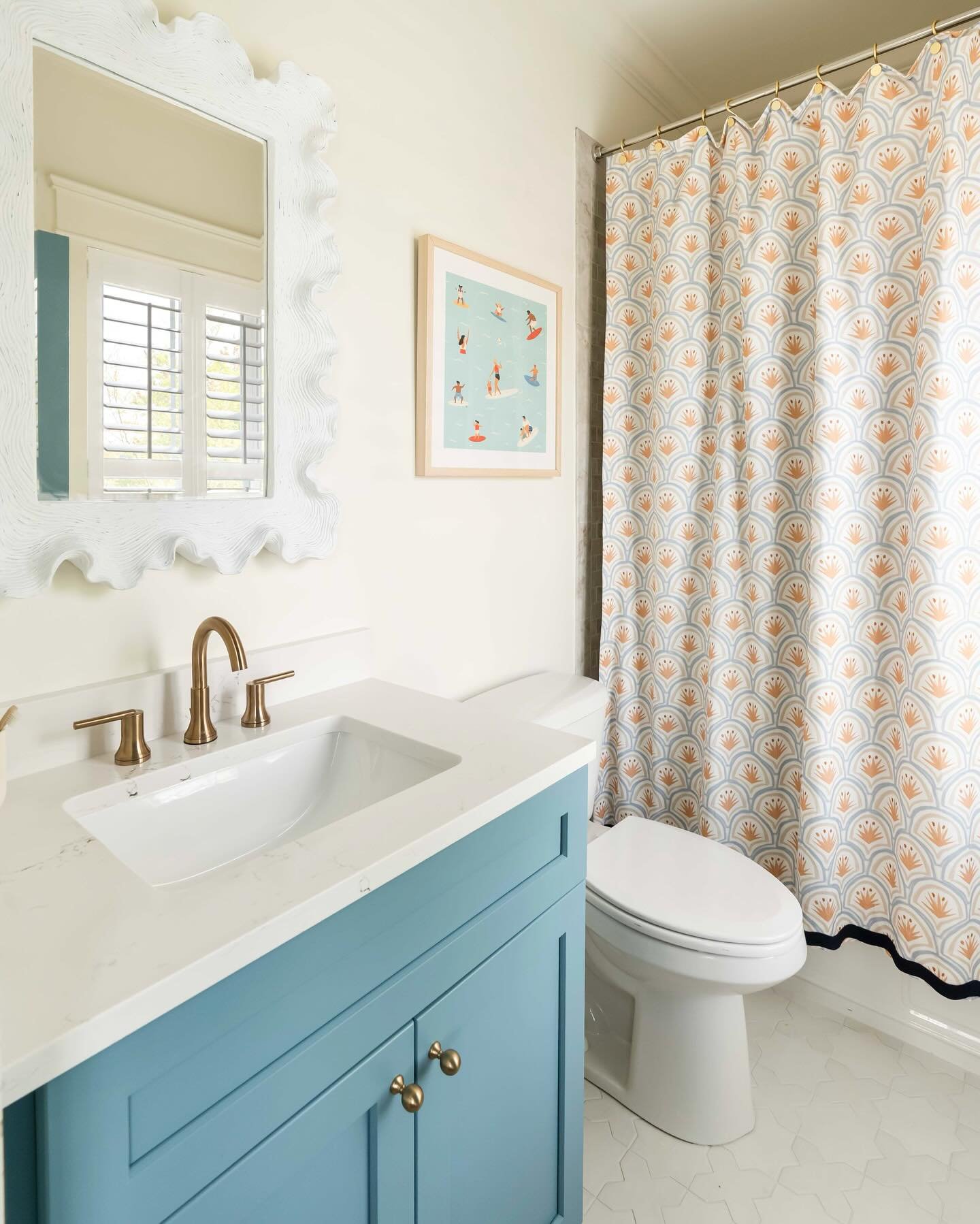 A white and light blue bathroom with a teal vanity, brass fixtures, and a shower curtain featuring a repeating orange and blue fan or scallop pattern.