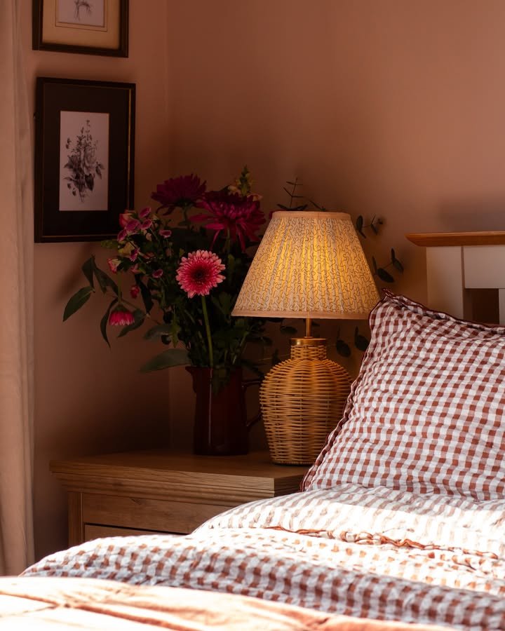A rattan or woven-based lamp with a patterned shade sits on a wooden nightstand beside a bouquet of flowers.