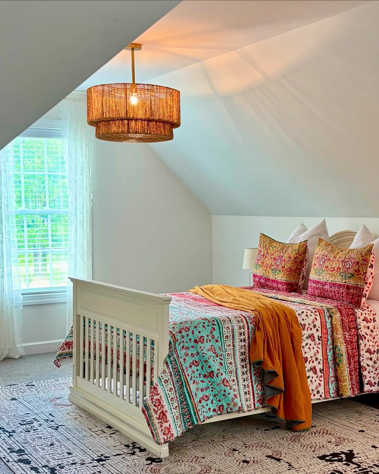 A two-tiered, woven pendant light with a textured, natural fiber design hangs from the slanted ceiling of an attic bedroom with a white bed and colorful patterned bedding.