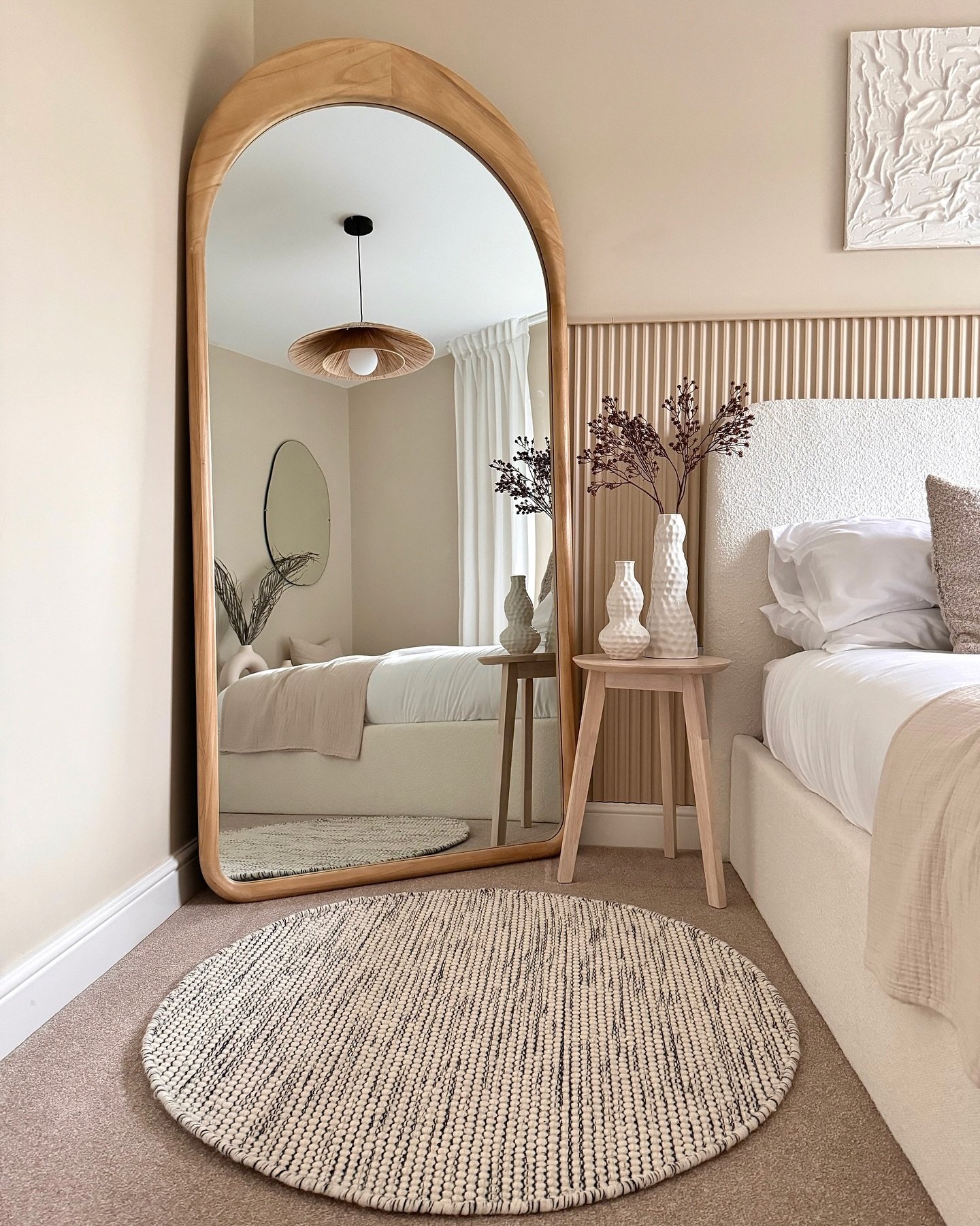 A tall, arched, natural wood-framed mirror leans in the corner of a neutral-toned bedroom, next to a bed with a textured headboard.