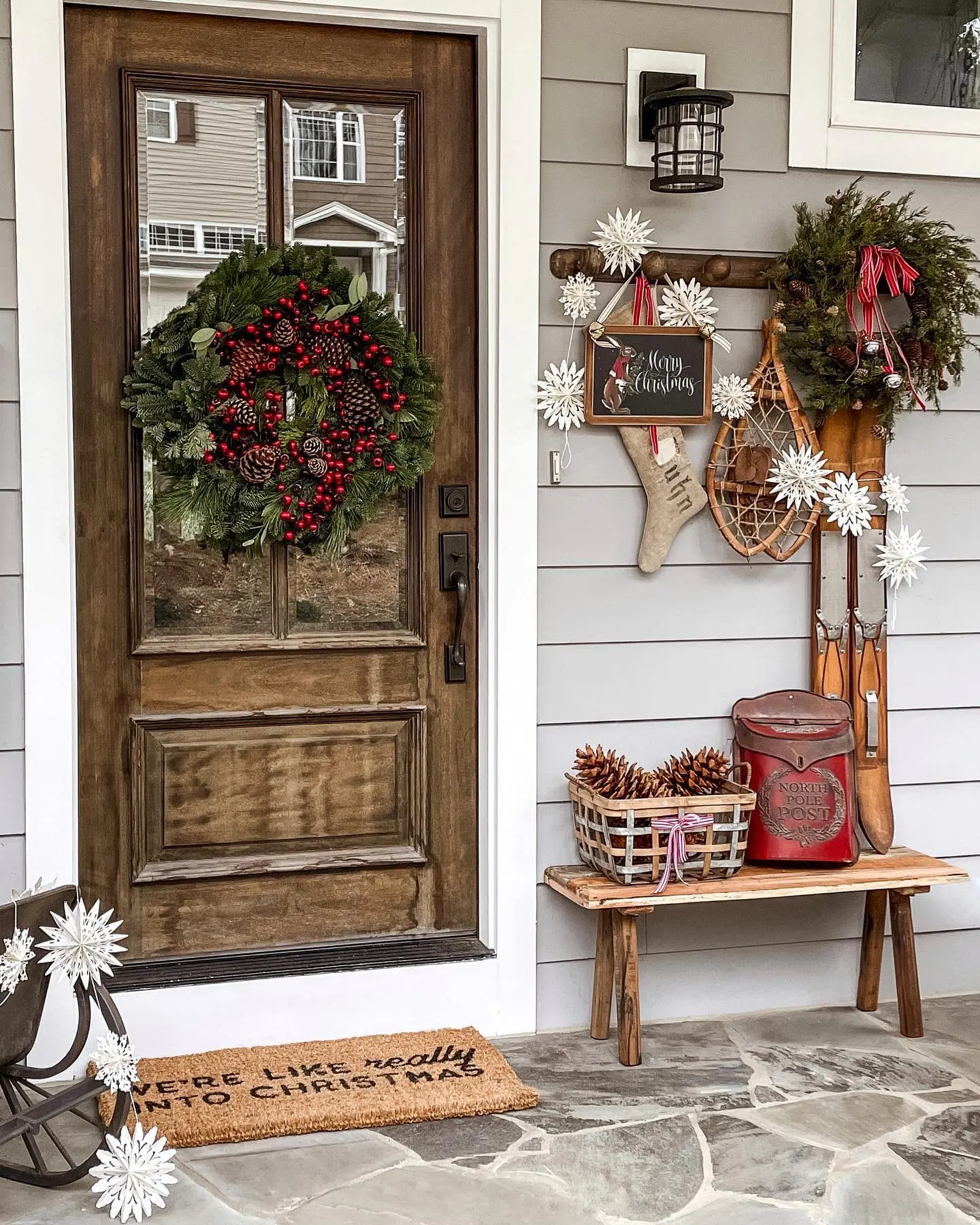 A cozy farmhouse Christmas porch with a rustic wooden bench, a vintage red "North Pole Post" mailbox, snowshoes, and a highly decorated wreath on a wood-grain door.