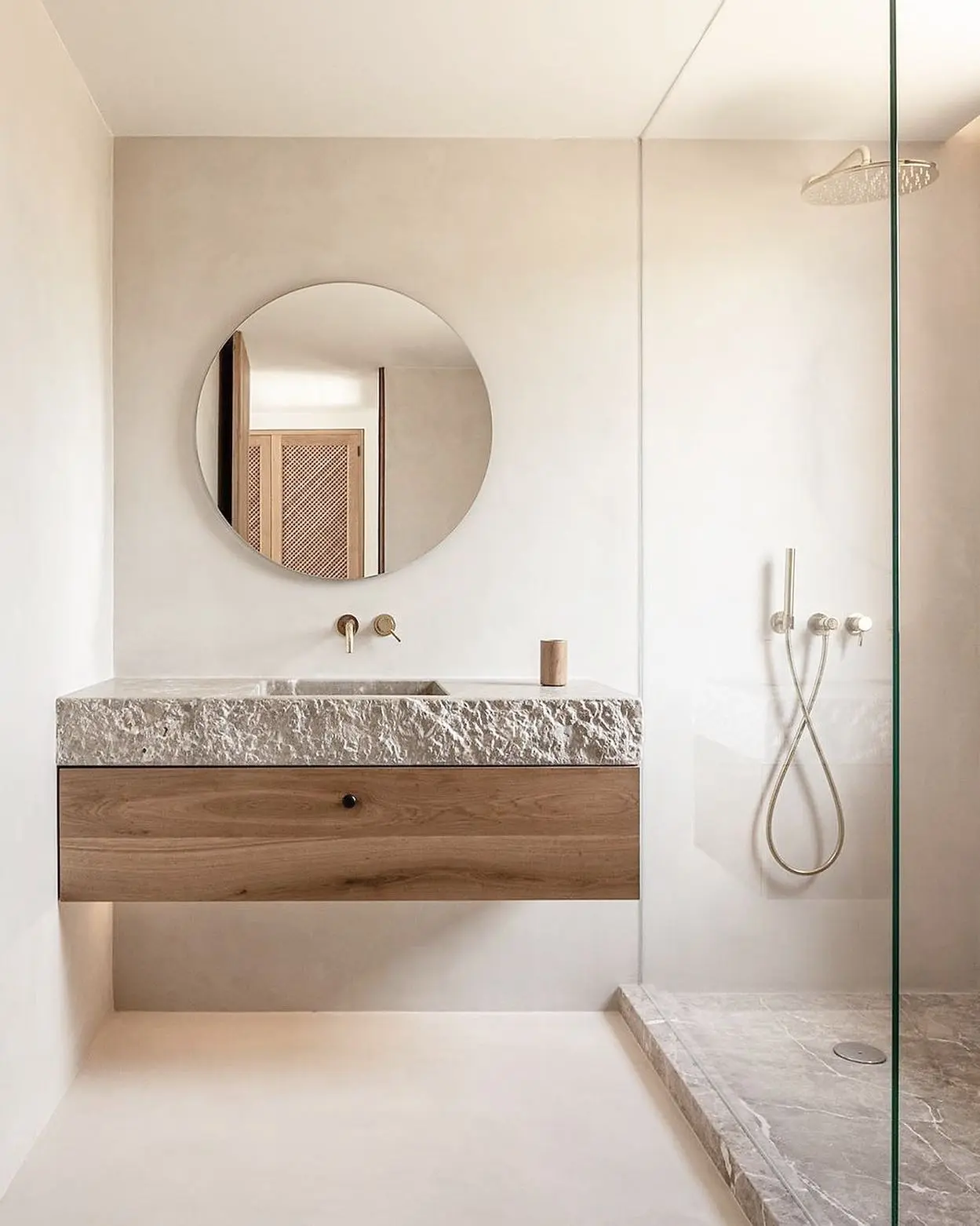 A floating bathroom vanity featuring a rustic, chiseled stone basin top resting on a light wood drawer with a small black knob. A round mirror hangs above it with a brushed gold faucet and fixtures, against a smooth, off-white wall.