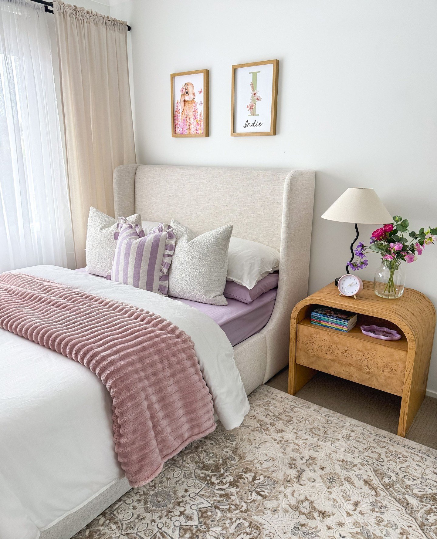 A cozy bedroom featuring a bed with a high, upholstered headboard, pillows in shades of lavender and white, a pink throw blanket, and a curved wooden nightstand with fresh flowers.