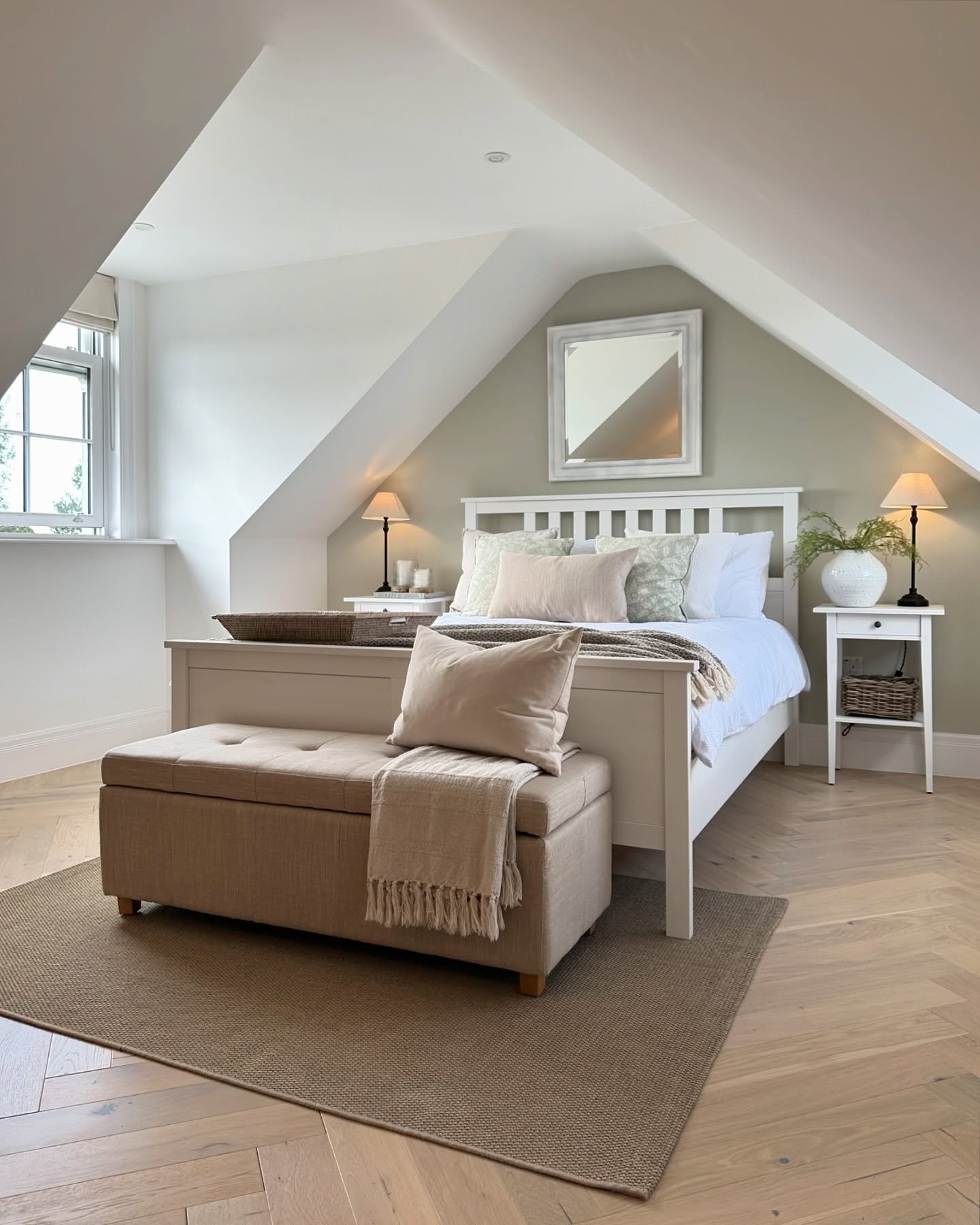 A bright attic bedroom with a white bed, beige upholstered bench at the foot, and light wood herringbone floors. The walls are a mix of white and soft sage green.