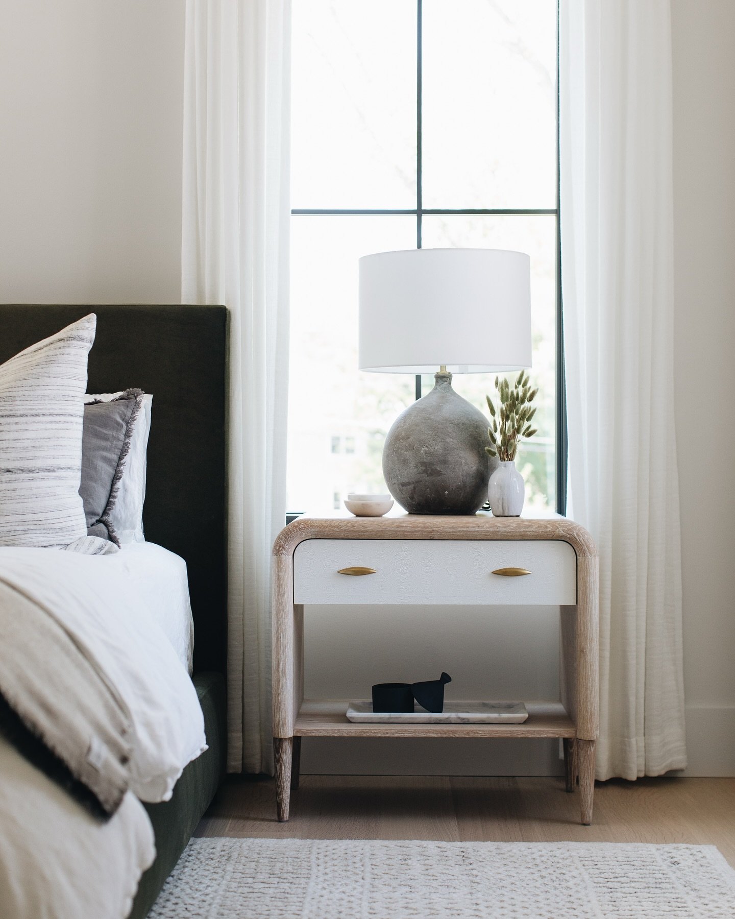 A two-tone nightstand with a light wood frame and white drawer front with gold pulls. The top holds a round, textured grey lamp, a small white vase with dried grasses, and a little bowl. The open shelf below contains a white tray with a decorative object.