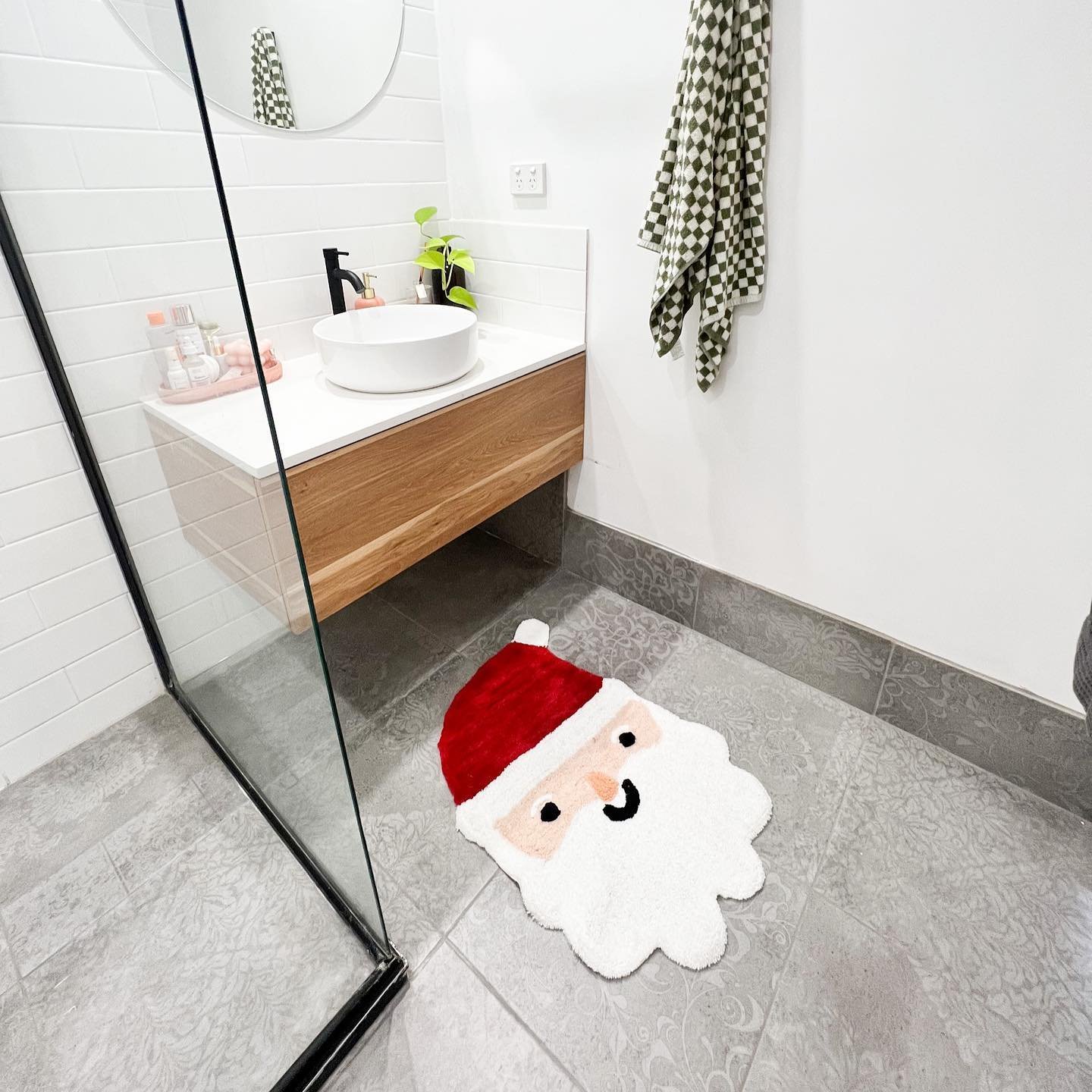 A modern white bathroom with a floating vanity and a playful bath mat shaped like Santa Claus's face on the floor.