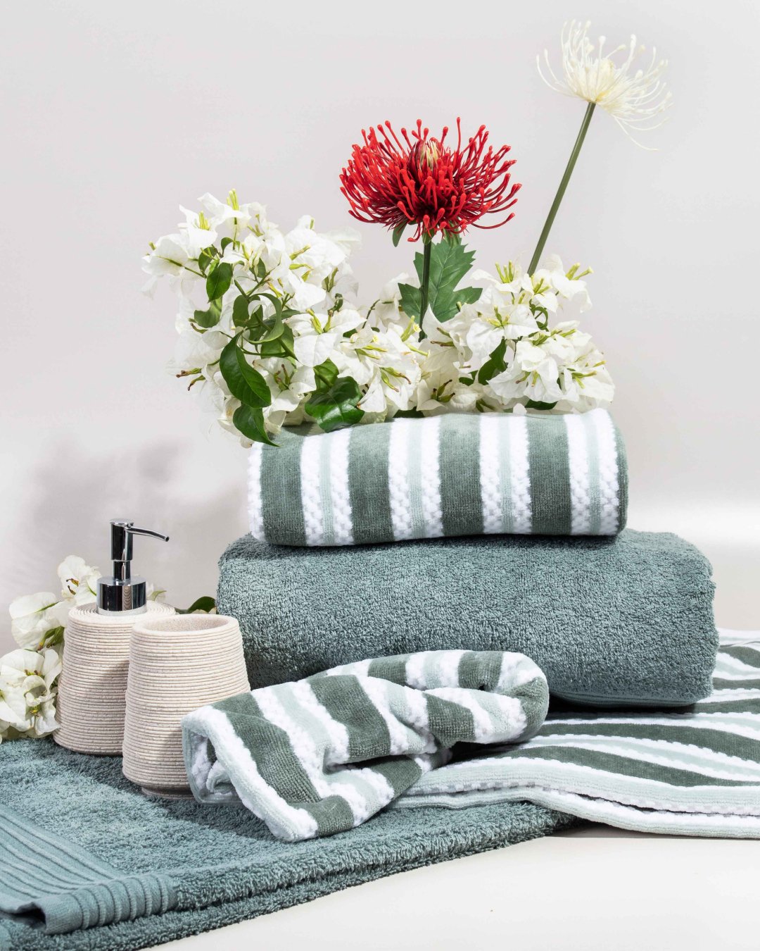 Stacked green and white striped towels and solid sage green towels, styled with ceramic accessories and flowers.