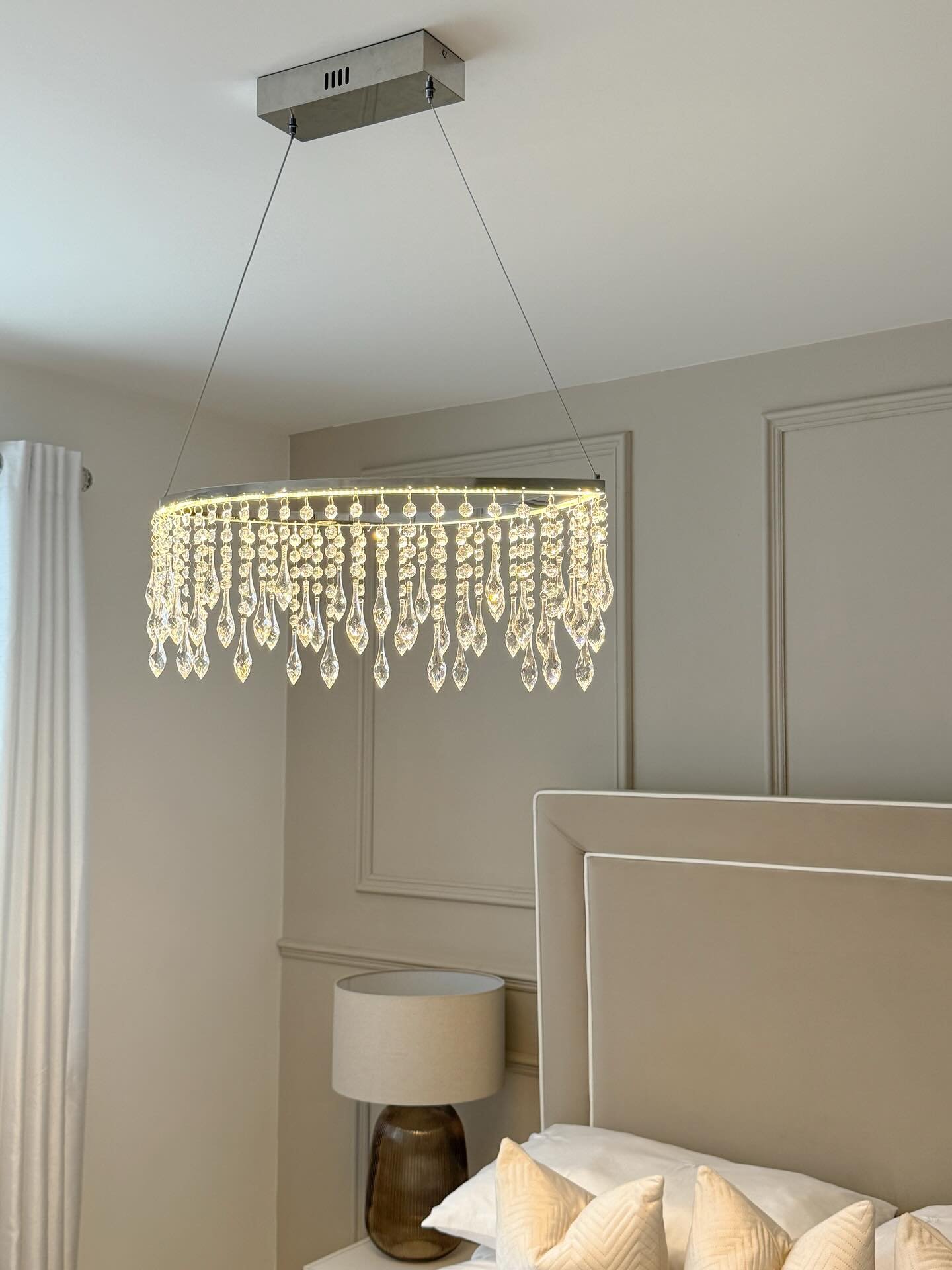 A contemporary rectangular chandelier with multiple rows of dangling clear crystal teardrops hangs from a silver base over a bed.