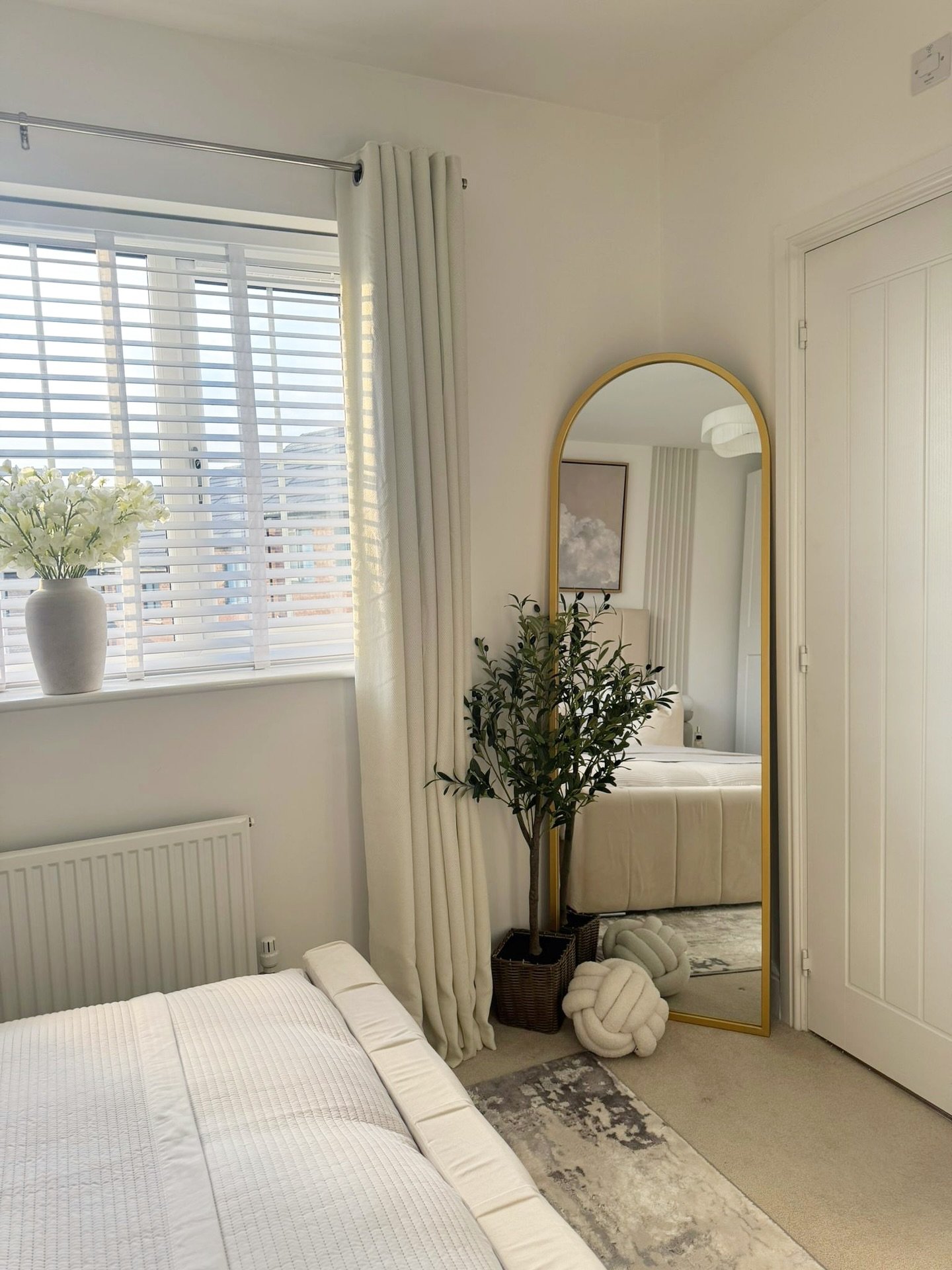 A tall, gold-framed arched mirror stands in the corner of a bright, neutral bedroom, next to a window with white blinds and curtains.