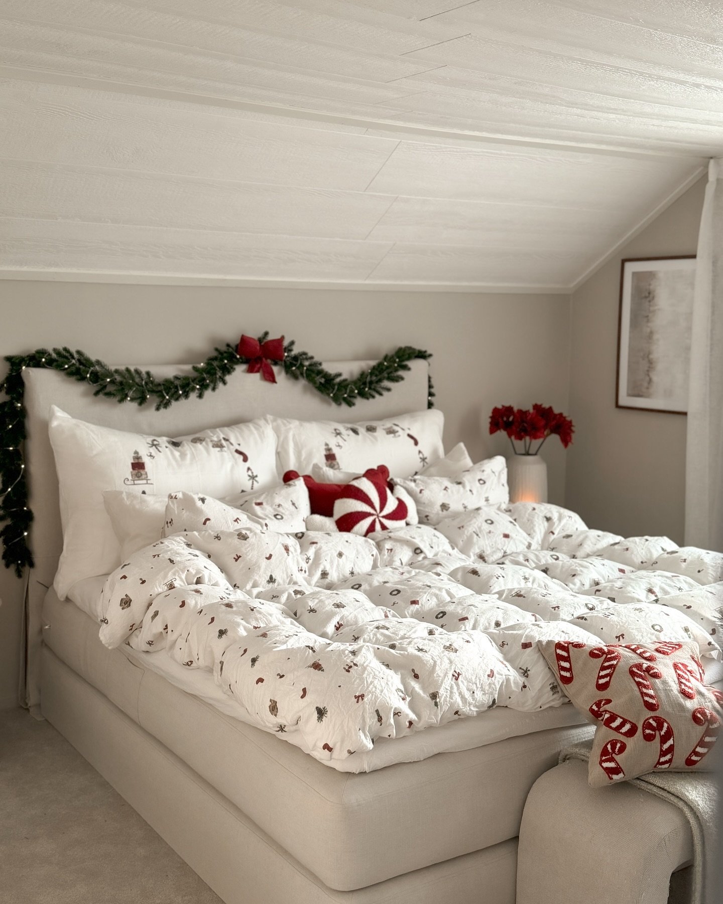  A bright bedroom with an angled ceiling features an all-white bed with a tiny Christmas pattern duvet, a garland draped across the headboard, and a burlap candy cane accent pillow.