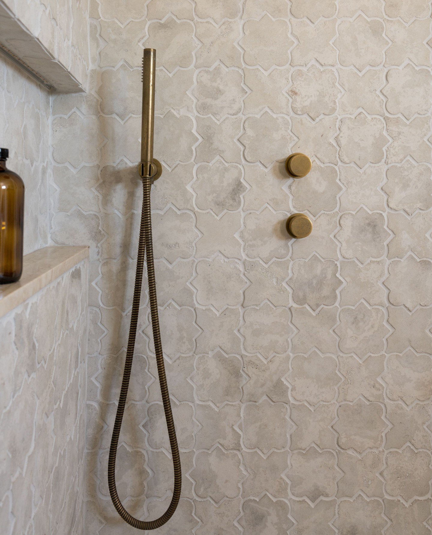 A close-up of a shower wall covered in light, sandy-colored, floral-shaped Moroccan tiles with dark grout and rustic brass fixtures.