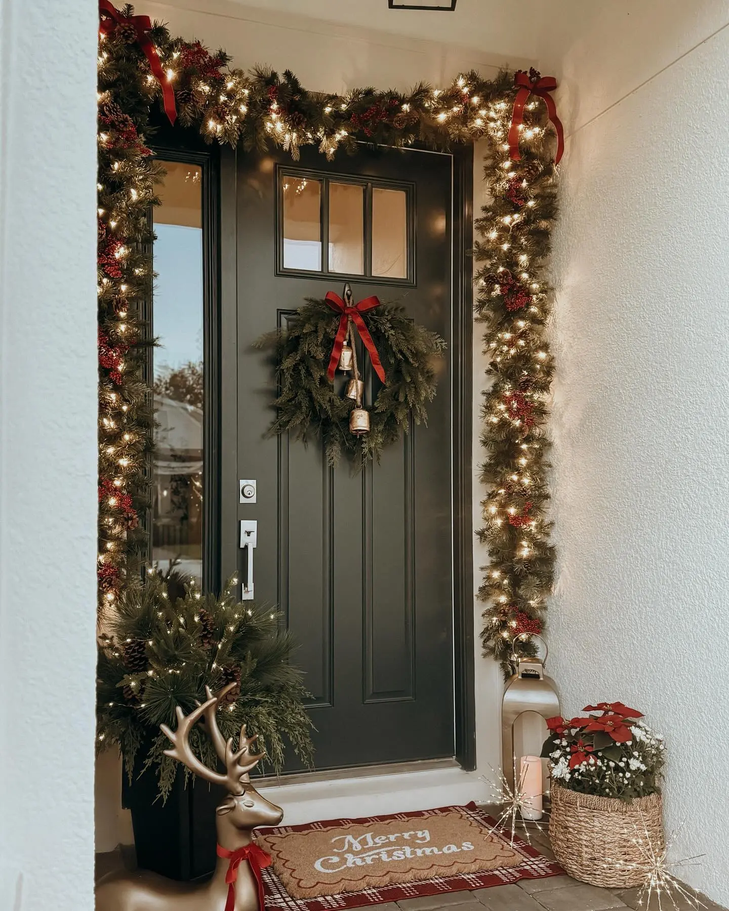 A luxurious Christmas porch featuring thick, heavily lit garland surrounding a dark front door, a golden reindeer statue, and a woven basket of poinsettias.
