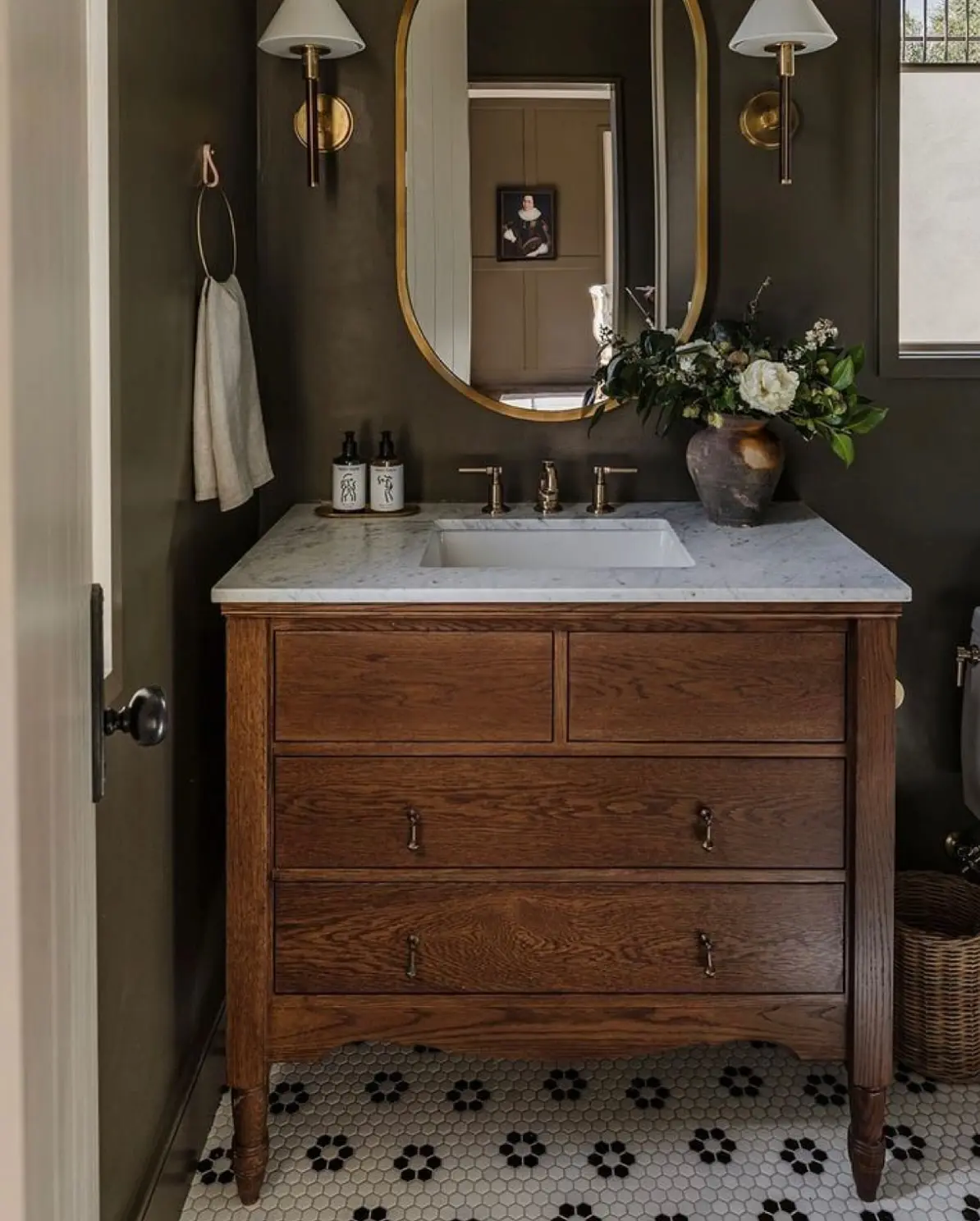 A traditional brown wood dresser converted into a bathroom vanity with a white marble countertop and a white undermount sink. It is paired with an oval gold-framed mirror and brass fixtures, against a dark olive-green wall.