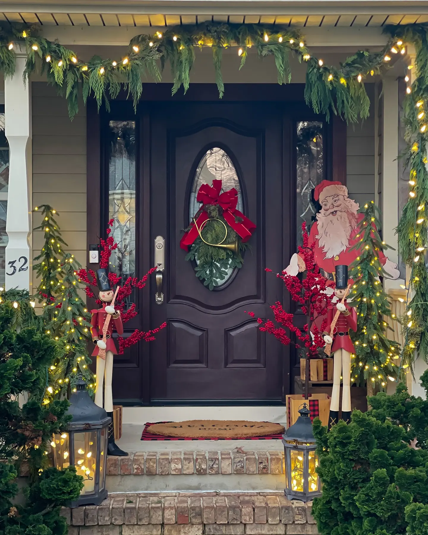 A festive Christmas porch featuring two tall wooden Nutcracker figures, a cardboard Santa cutout, and illuminated evergreen garland draped across a dark wood door.
