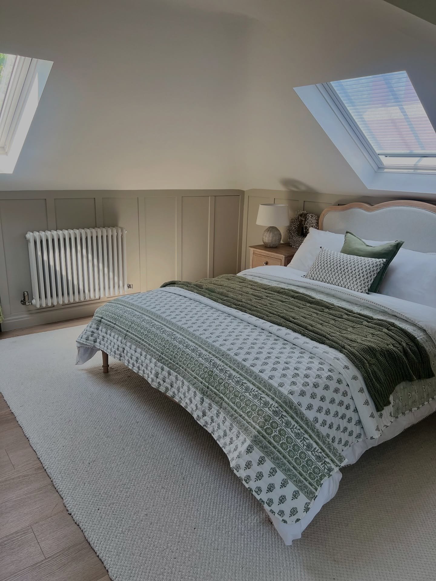 A cozy loft bedroom with sloped ceilings, two skylights, and soft green paneled walls. The bed is dressed in white bedding with a green patterned quilt.
