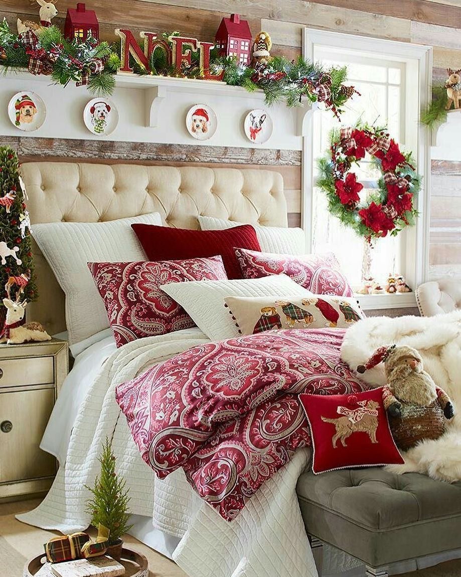 A cozy bedroom with a wooden shiplap wall, featuring a bed with deep red paisley bedding, a tufted headboard, and a shelf adorned with miniature red houses, a "NOEL" sign, and dog-themed Christmas plates.