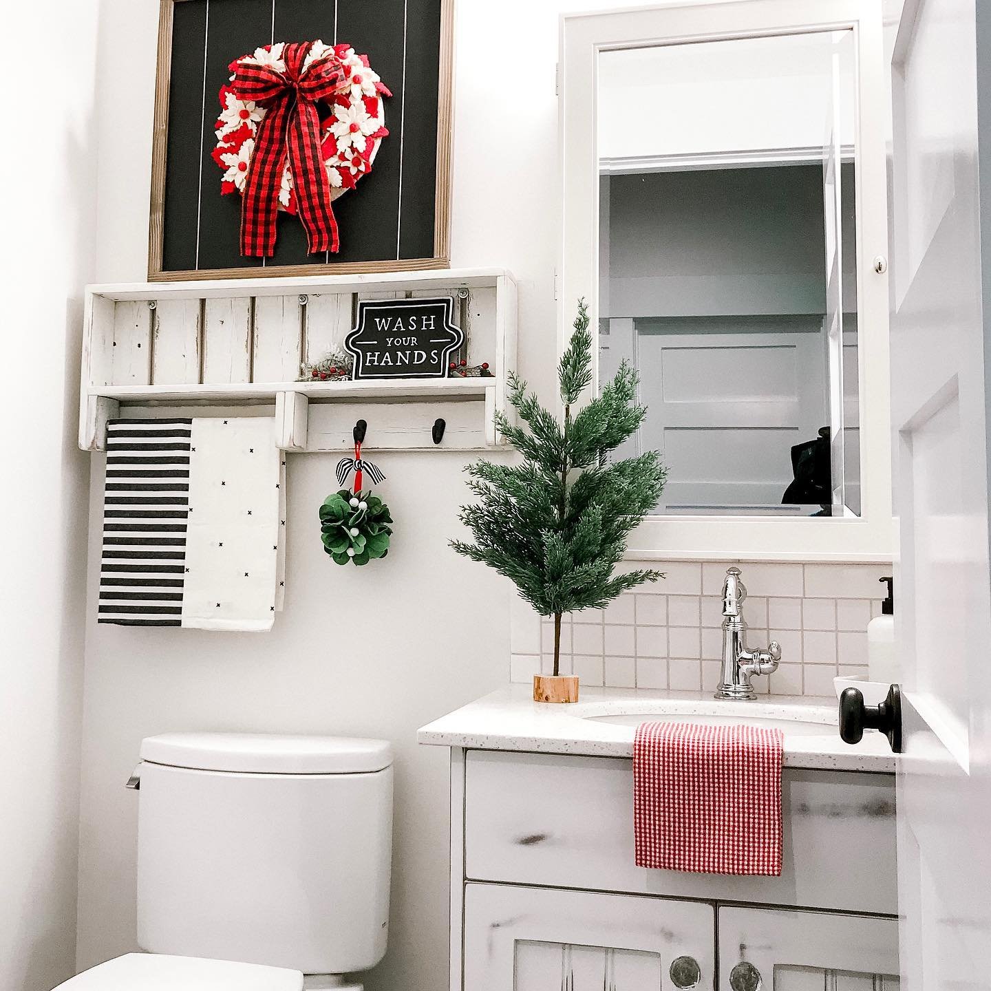 A small white farmhouse bathroom featuring buffalo plaid accents, a red floral wreath, and a tabletop Christmas tree next to the sink.