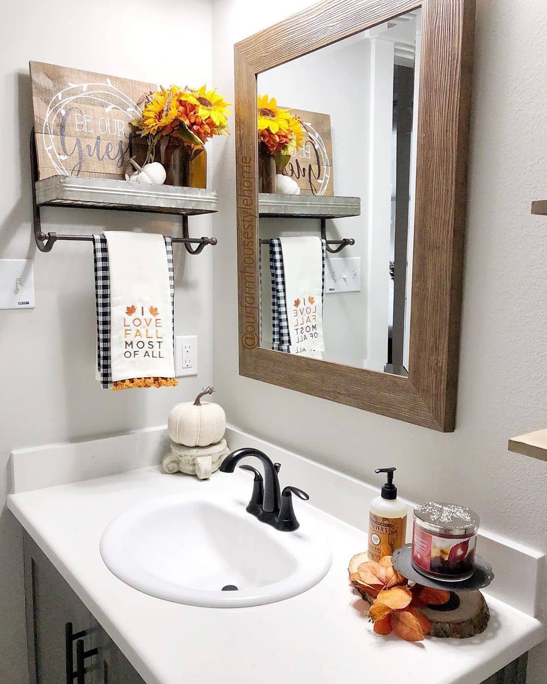 A white bathroom sink with a wooden mirror, rustic wall shelf, sunflowers, and fall-themed hand towels.