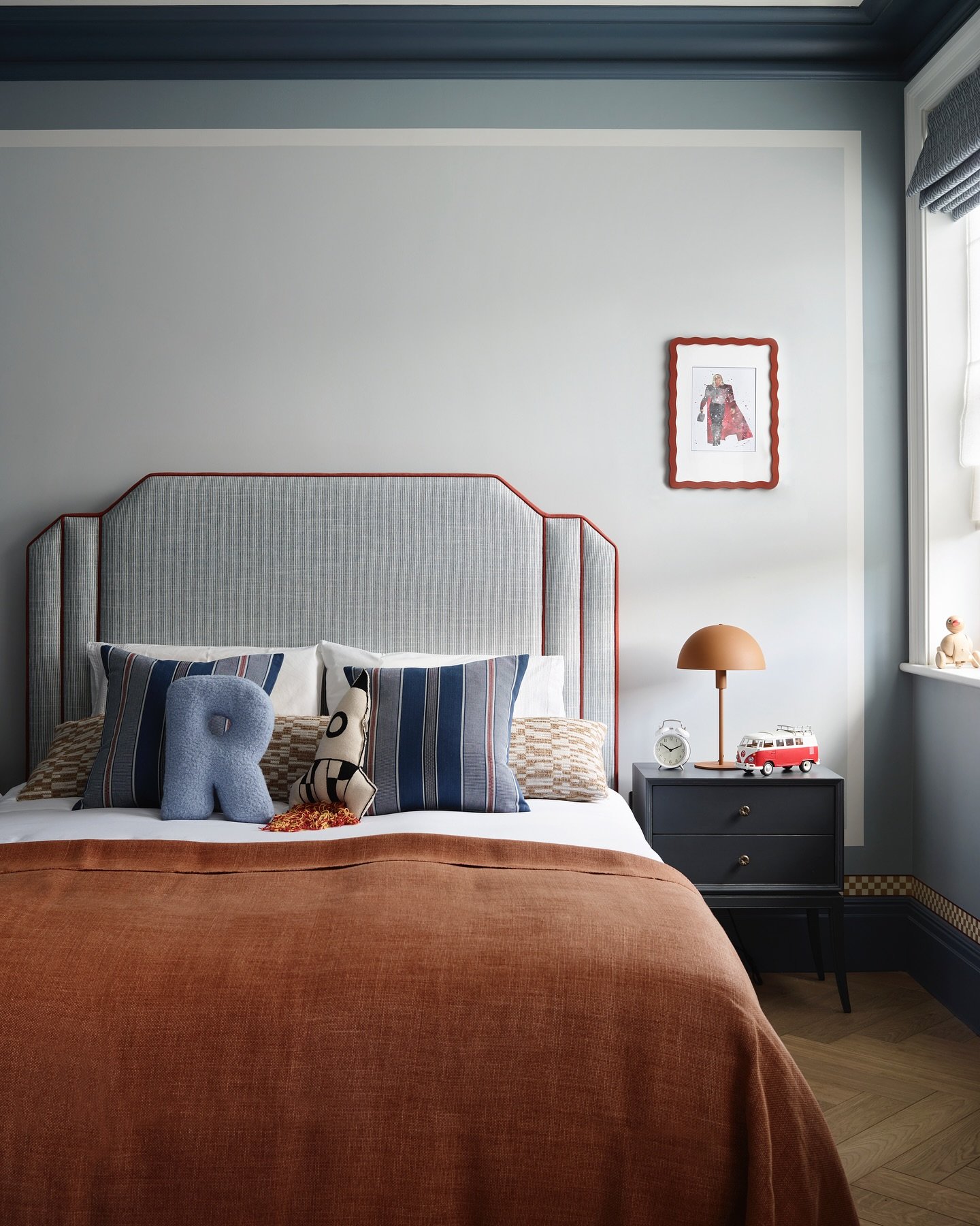 A child's or teen's bedroom featuring a bed with a striped blue headboard, a rust-colored blanket, a 'R' shaped pillow, and a small picture frame on the wall.