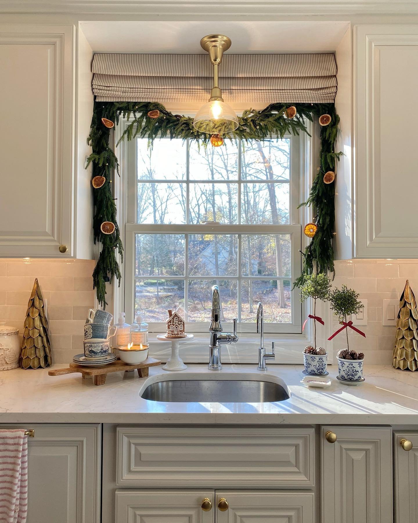 A bright kitchen window decorated with a pine garland accented by dried orange slices and string lights. The sink area features a small gingerbread house, candles, blue and white patterned china, and potted evergreen topiaries.