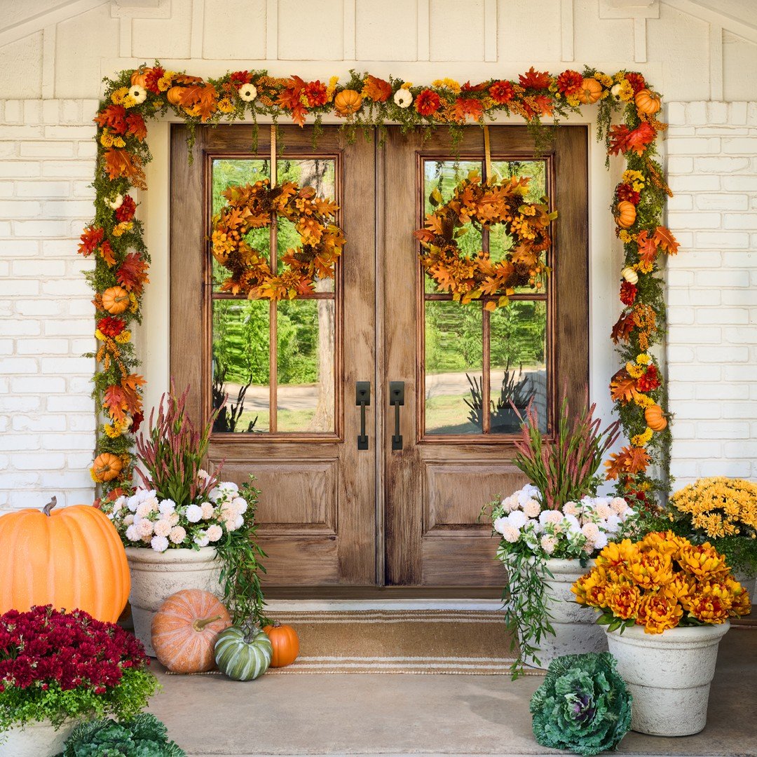 A vibrant fall porch decorated with pumpkins, planters of mums and cabbages, and a rich garland of leaves and mini pumpkins framing the wooden double doors.