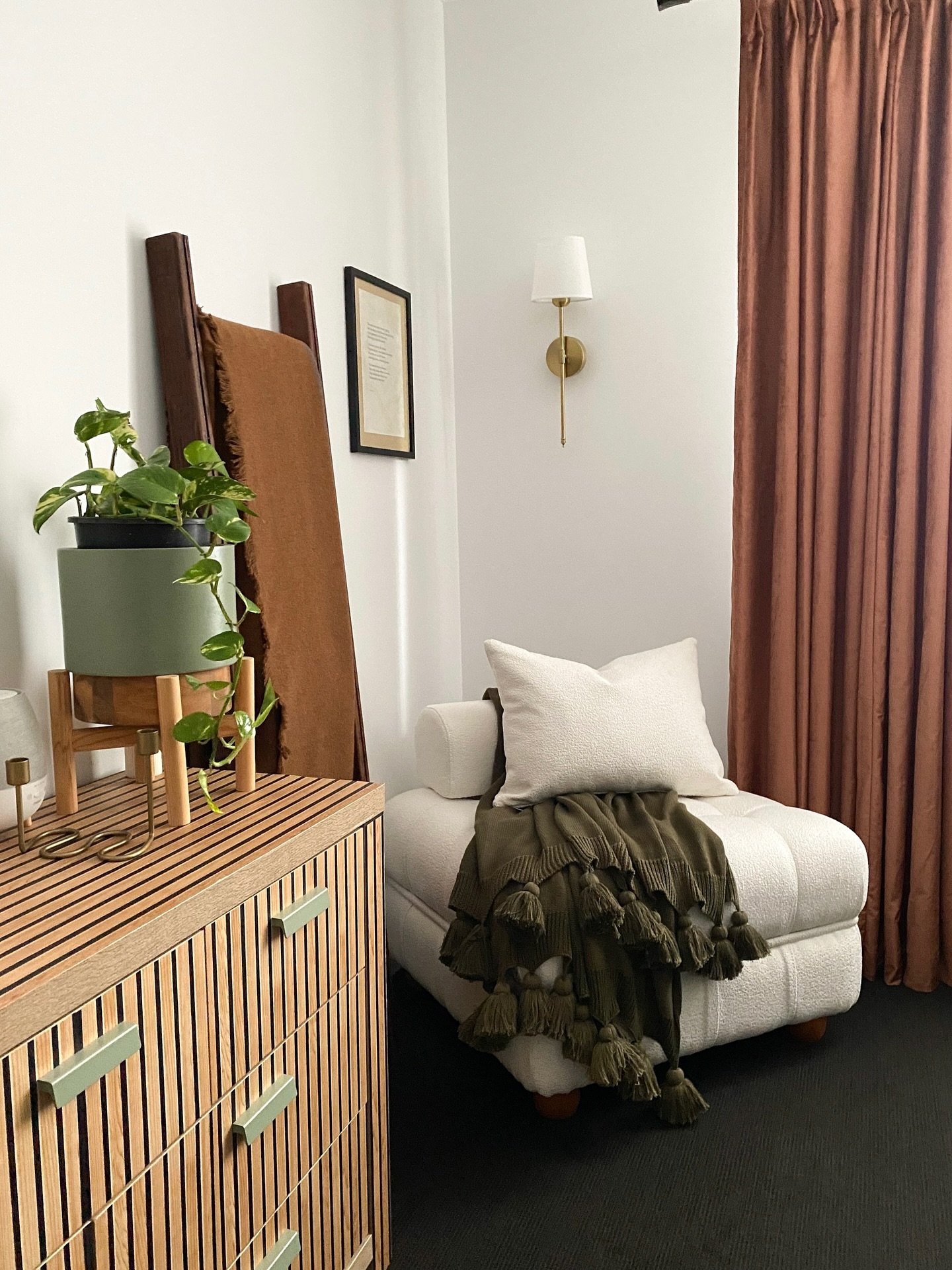 A small cream-colored armchair with an olive-green tassel blanket sits in a bedroom corner, next to a wooden chest of drawers and a potted plant.