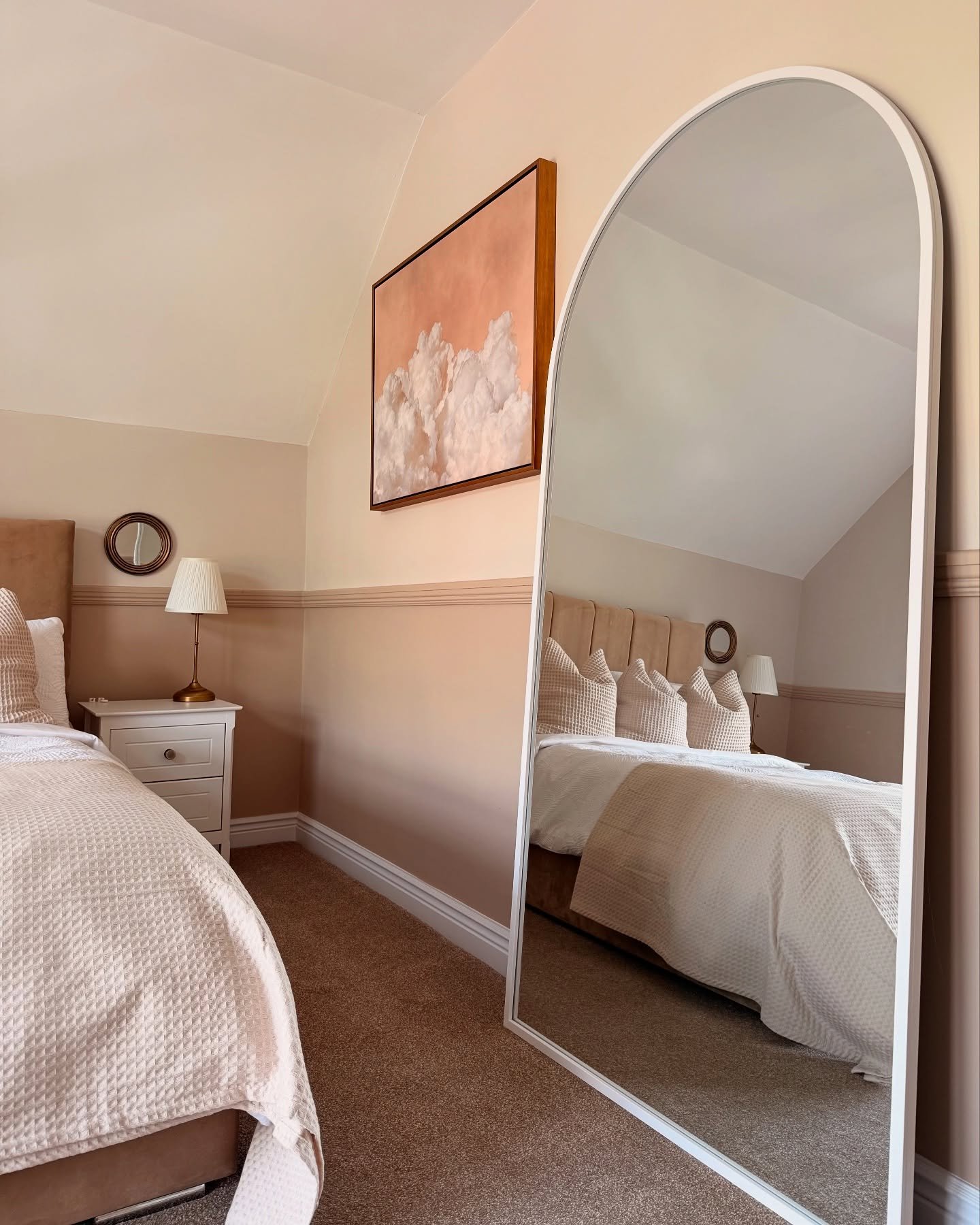 A tall, white-framed arched mirror leans in a bedroom with a two-toned wall, reflecting a bed with neutral-colored pillows and a textured blanket.