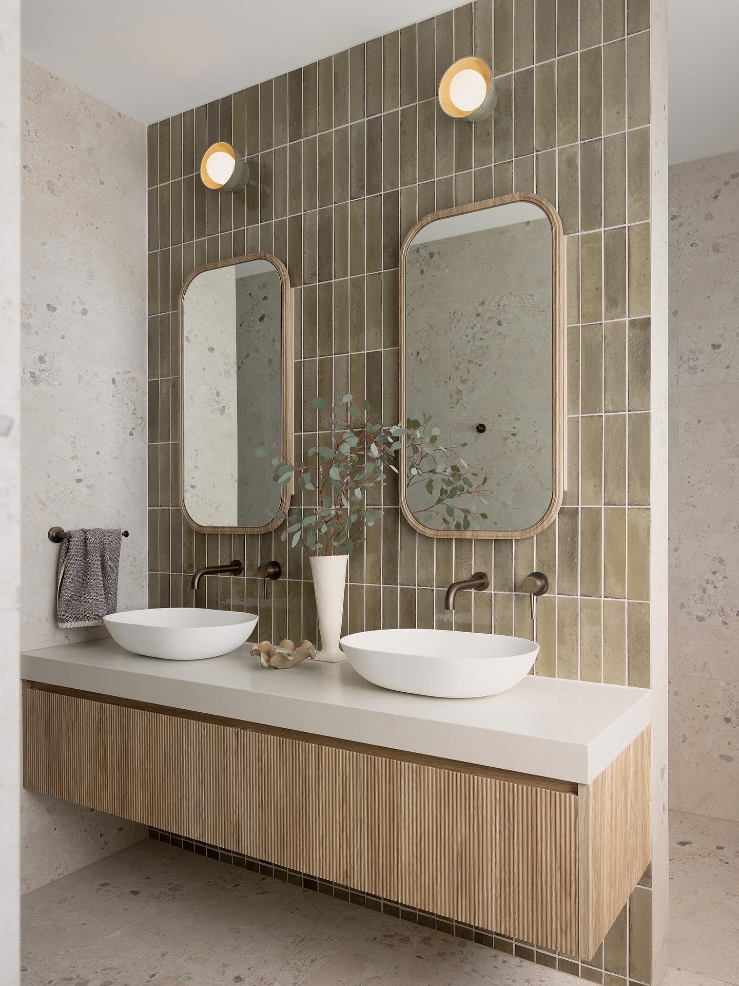 A double vanity in a modern bathroom with a light wood reeded cabinet, white vessel sinks, and a feature wall with vertical moss-green subway tiles.