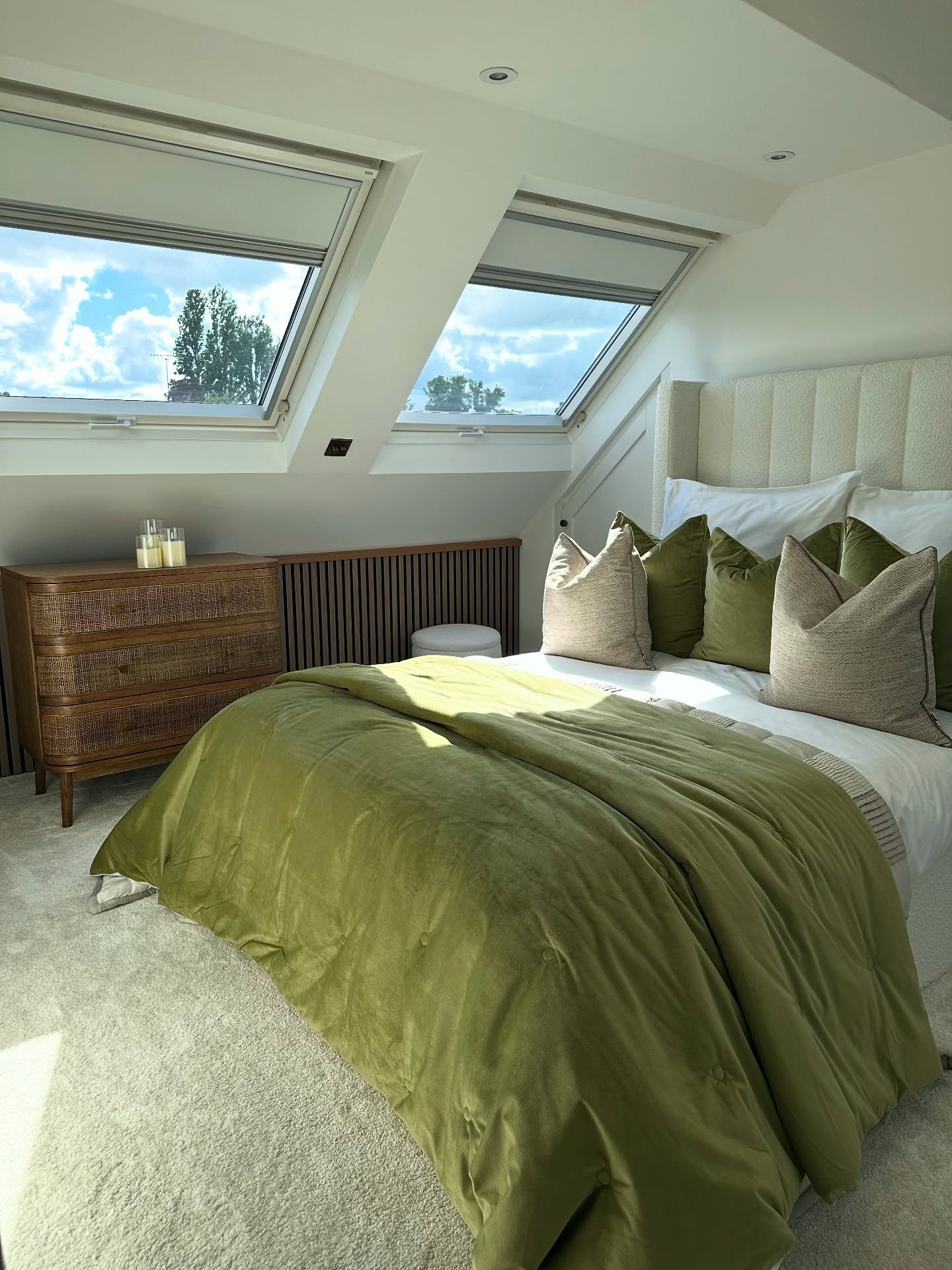 A modern loft bedroom with two skylights, a creamy white and olive green color palette, a plush headboard, and a wooden dresser with a textured, slatted front.