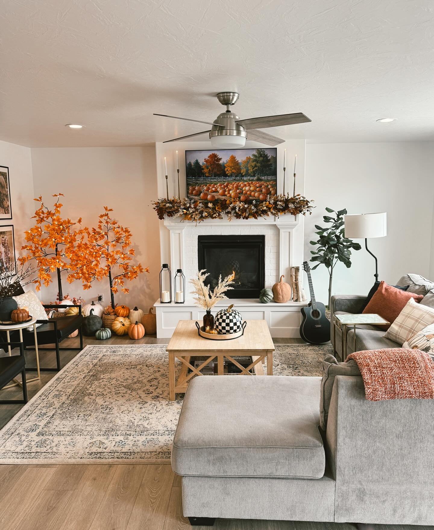 Spacious living room with a white brick fireplace, a pumpkin patch painting, orange artificial trees, and a gray sectional sofa.