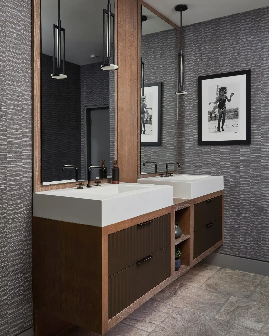 A floating double vanity with a warm wood frame and ribbed dark brown drawer fronts. The white sinks are integrated into the countertop, with black faucets, against a dark, patterned wallpaper and beneath two long black pendant lights.