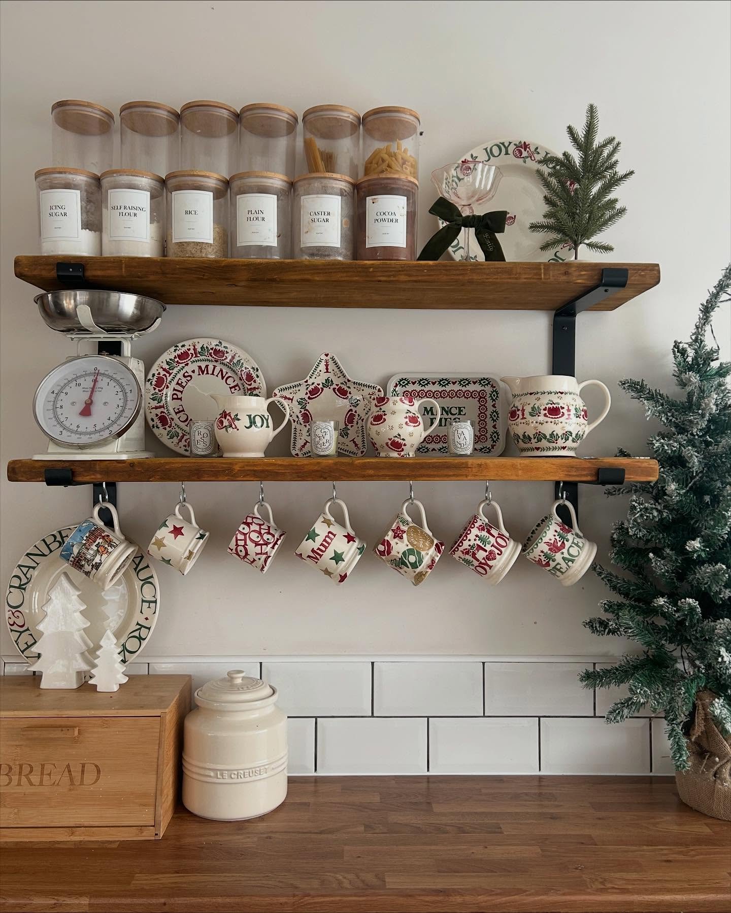 Kitchen open shelving styled for Christmas, featuring labeled canisters, a collection of vintage-style red and green holiday plates and mugs hanging from hooks, and a small potted Christmas tree on the shelf and floor.