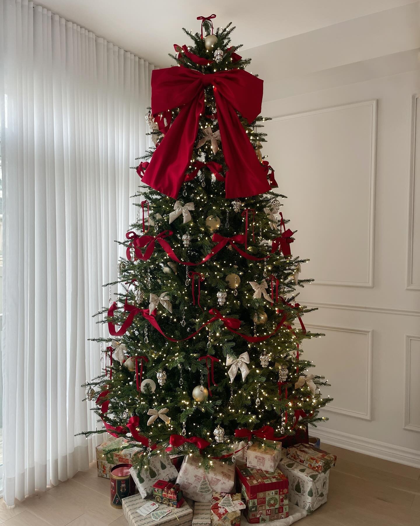 A Christmas tree heavily decorated with a dramatic oversized red velvet bow at the top, smaller bows, and long, swirling red ribbon garland.