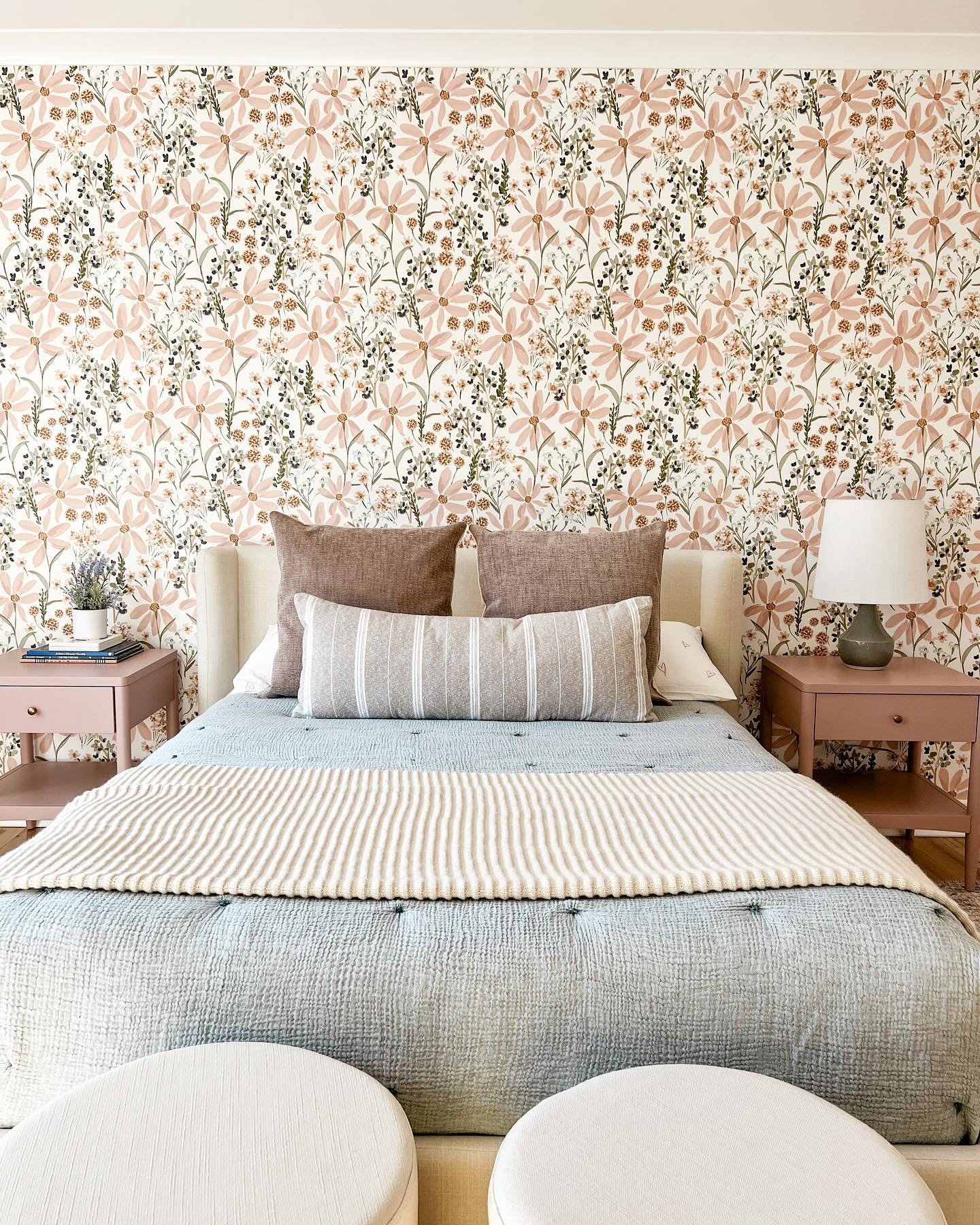 A bedroom with a prominent floral wallpaper in pink and brown, a bed with a blue quilt and beige throw, and matching pink nightstands on either side.