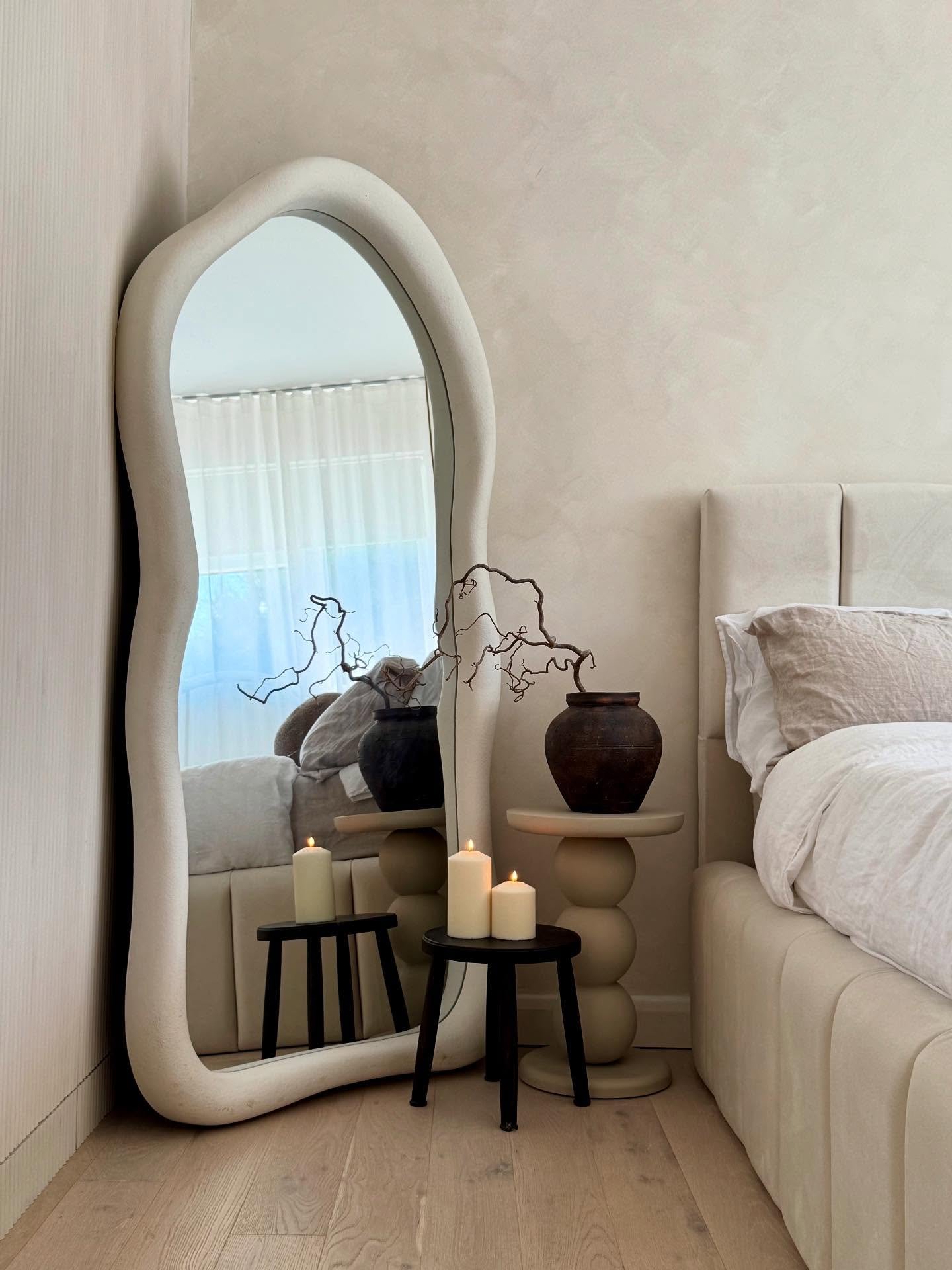 An irregularly shaped, cream-colored wavy mirror leans in a bedroom corner, reflecting a bed and two black stools with white candles.