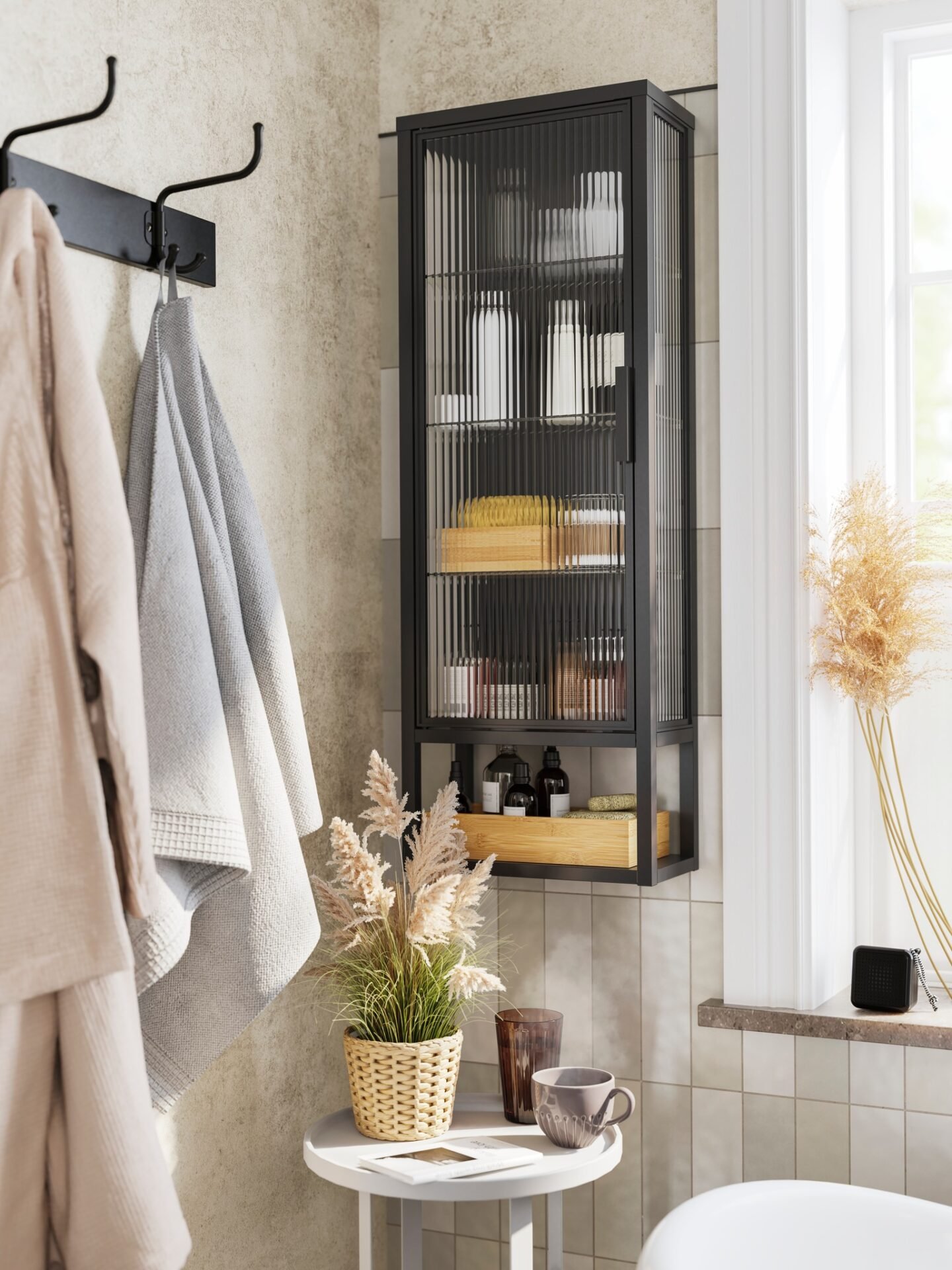 Black wall-mounted bathroom cabinet with reeded or fluted glass doors and two internal shelves, mounted next to a window and a black coat rack with towels.